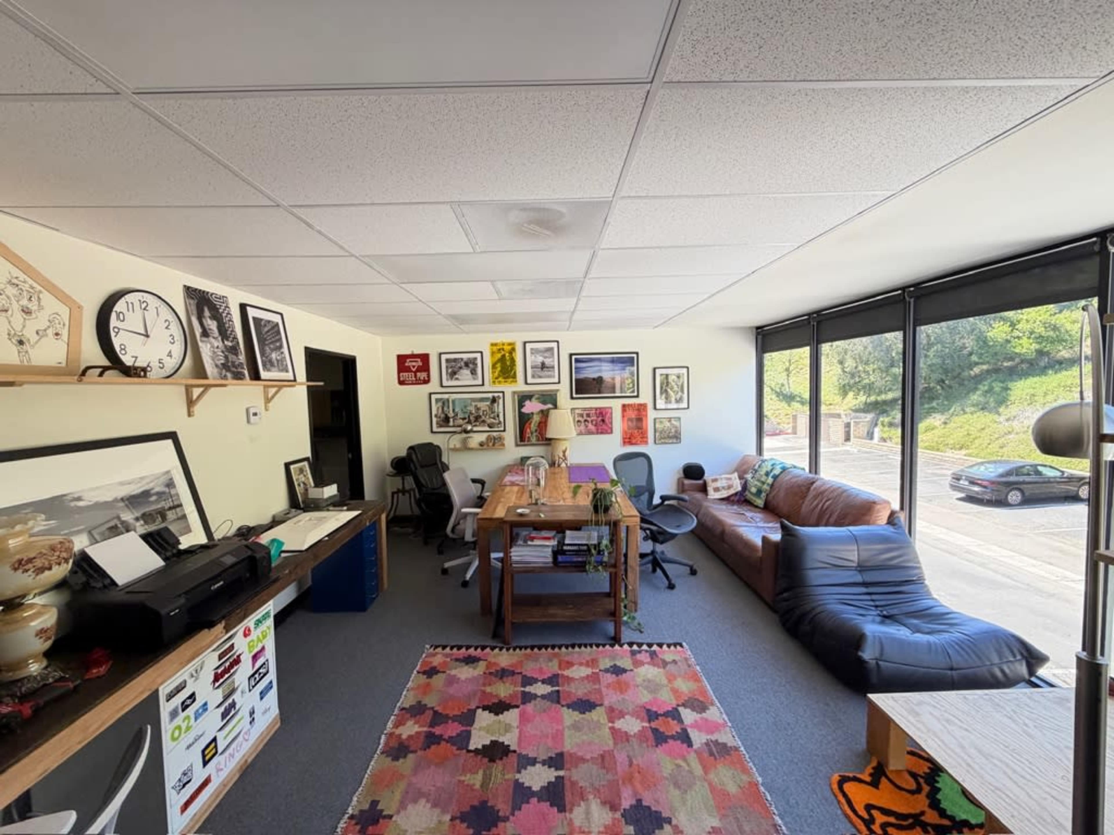 The image shows an office space with various artworks on the walls, a long wooden table, several chairs, and large windows allowing natural light to enter.