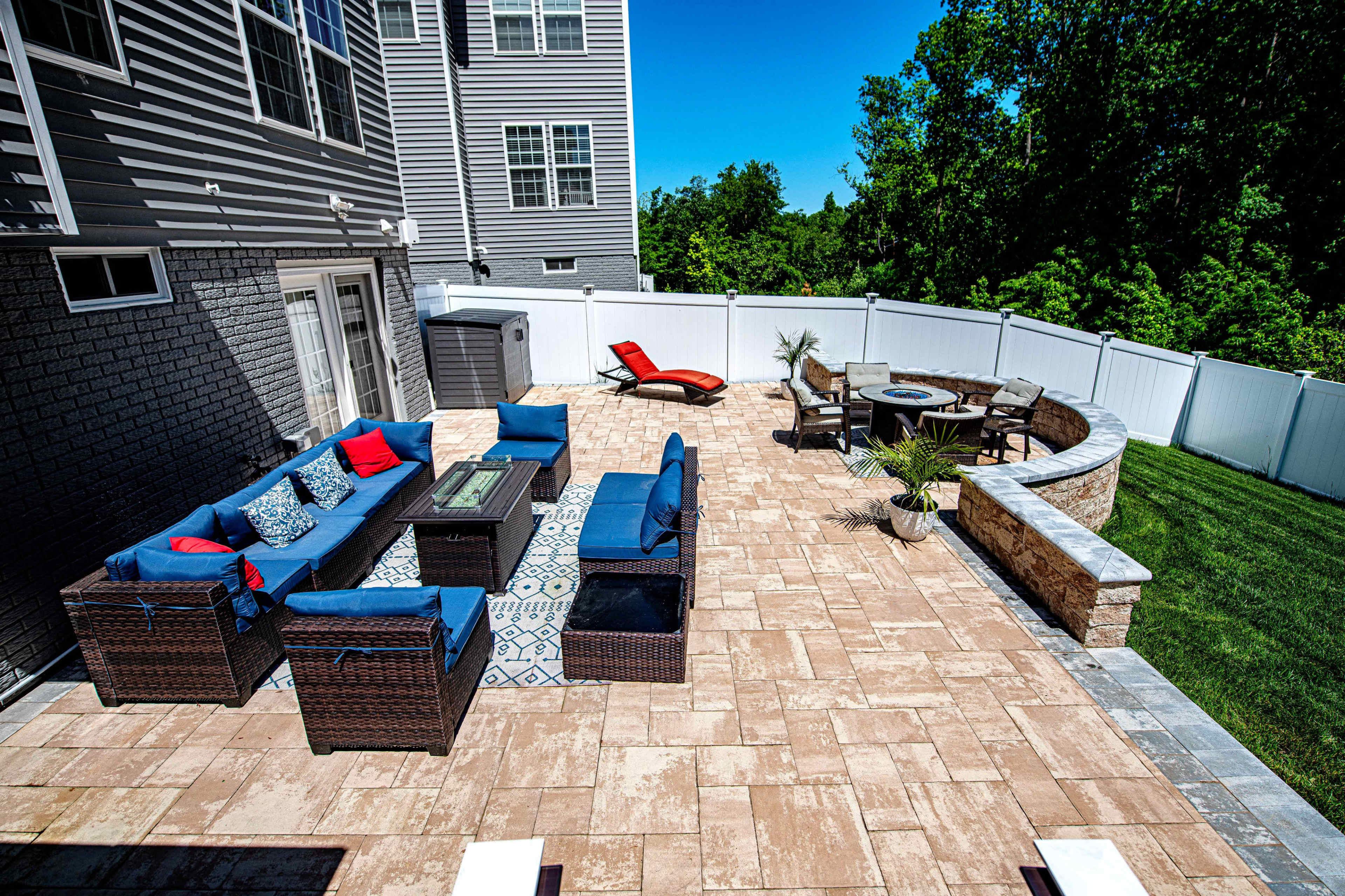 A spacious outdoor patio area featuring a sectional seating arrangement, a circular dining table, and a lounge chair, all surrounded by a grassy yard and a white fence.