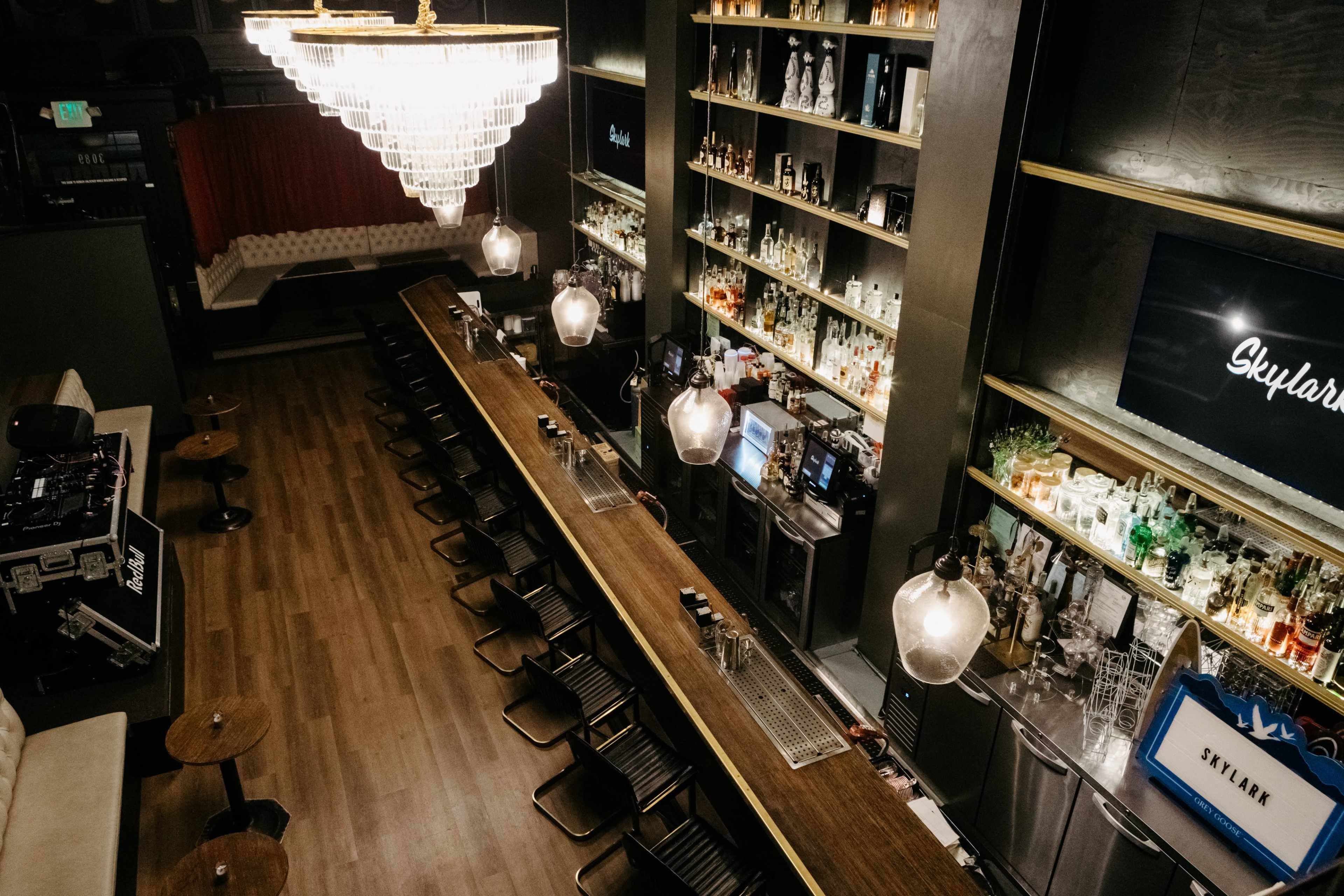 The image shows a modern bar interior with a long wooden counter, shelves lined with various bottles, and pendant lighting hanging above the bar.