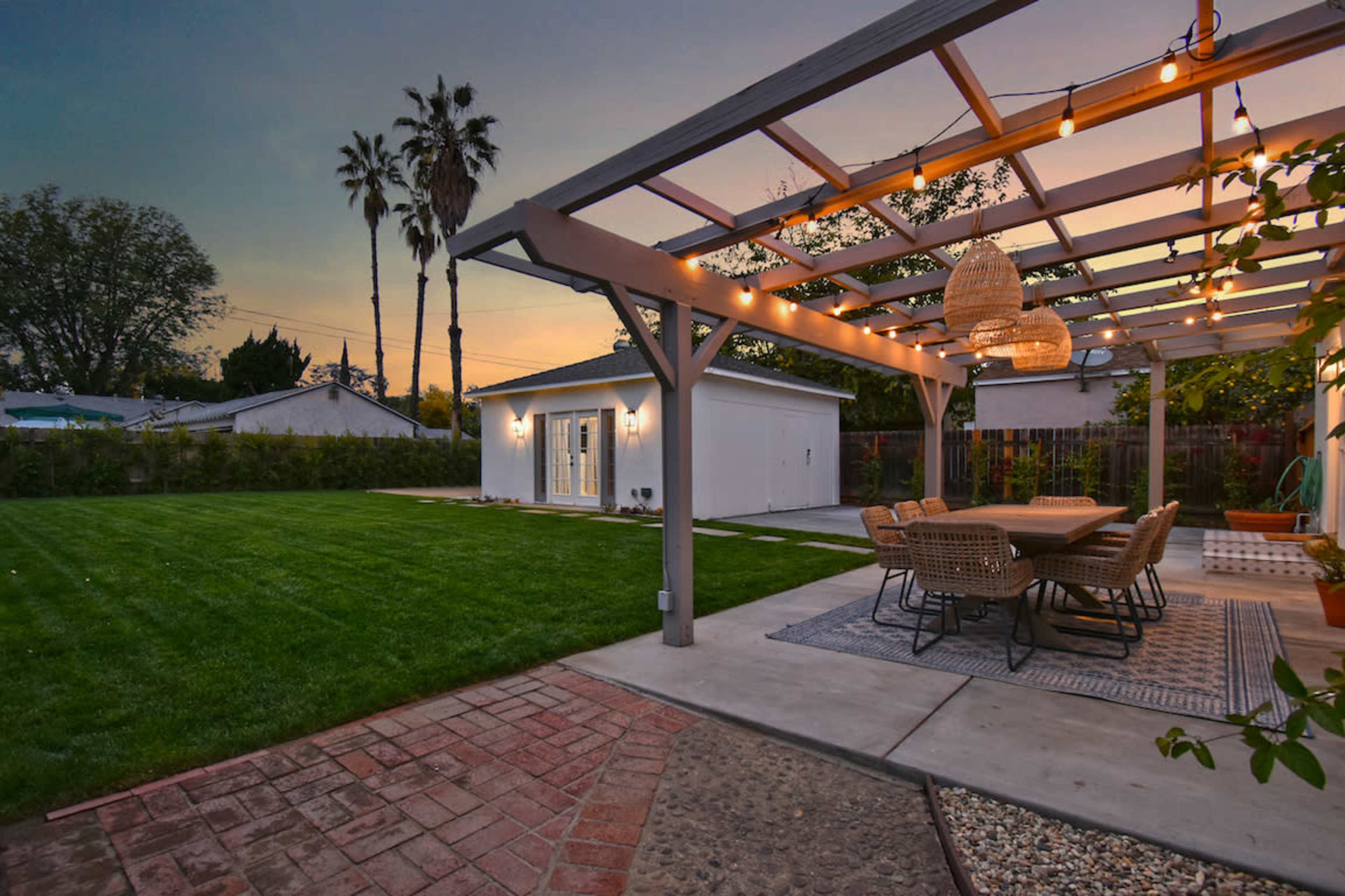The image shows a backyard setting with a pergola adorned with string lights, a dining area, and a white storage shed against a twilight sky.