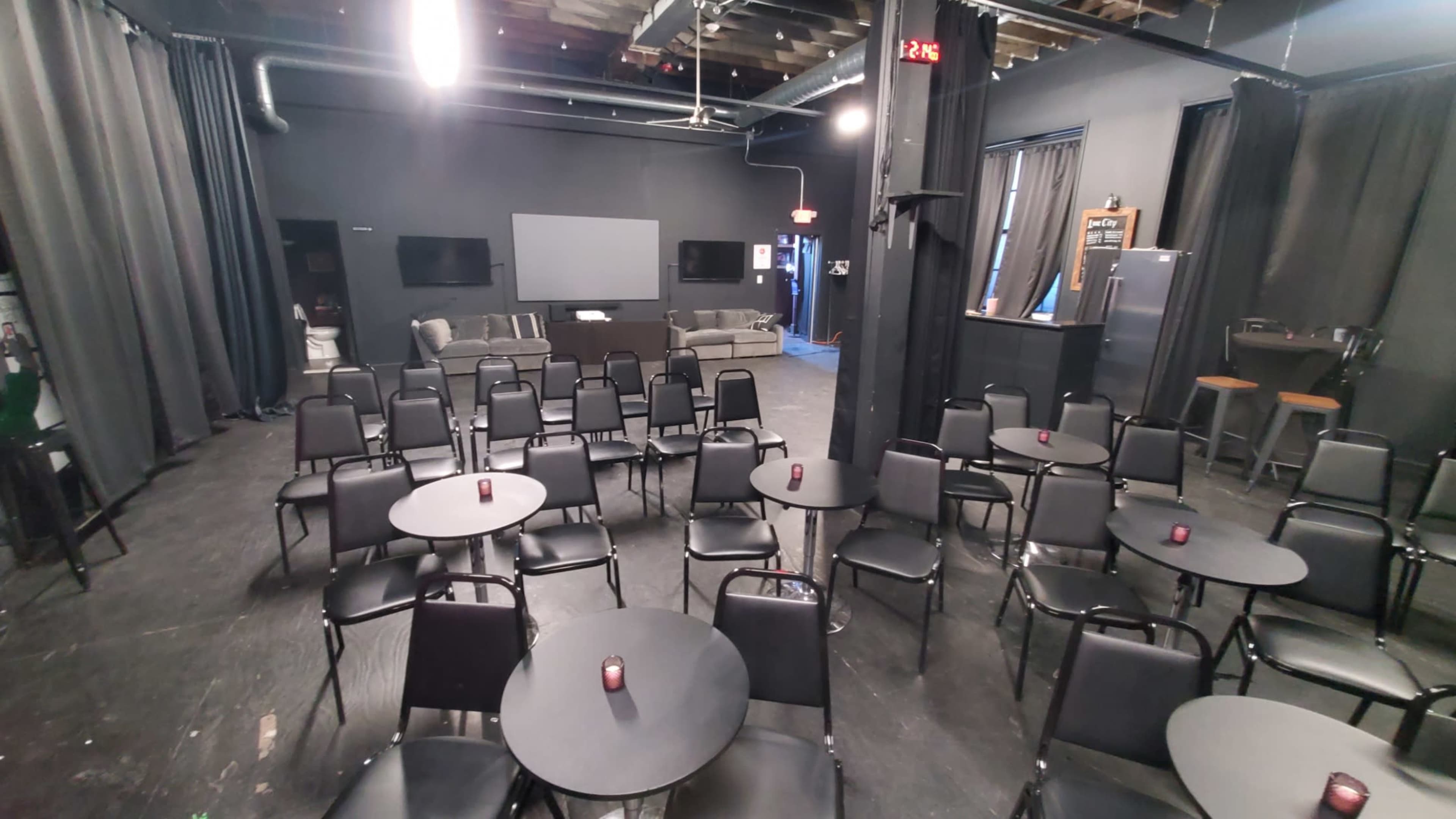 The image shows a dimly lit performance space with black chairs arranged around small tables facing a blank screen and a couch area in the background.