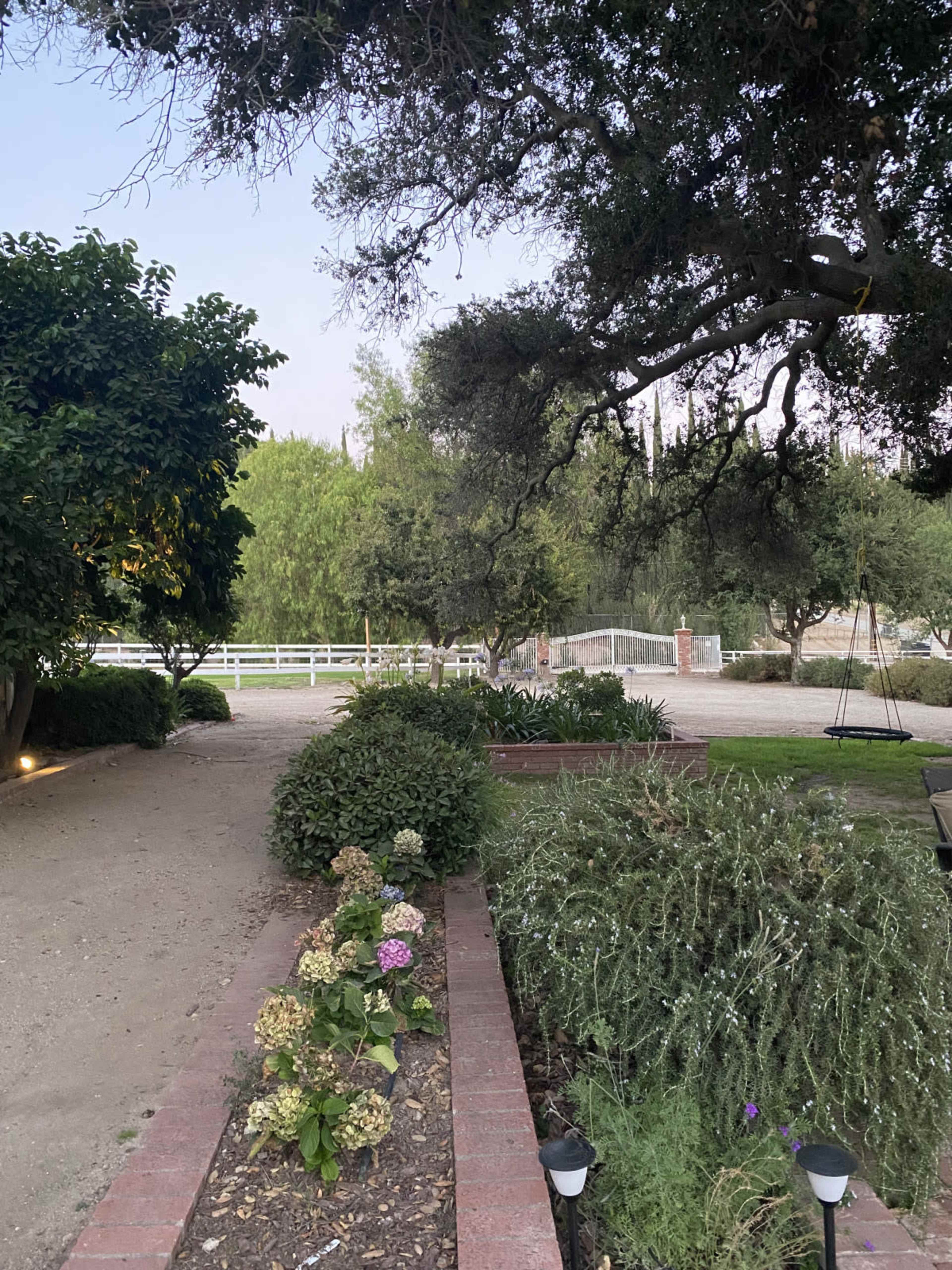 A landscaped path with blooming flowers leads to a grassy area enclosed by trees and a fence.