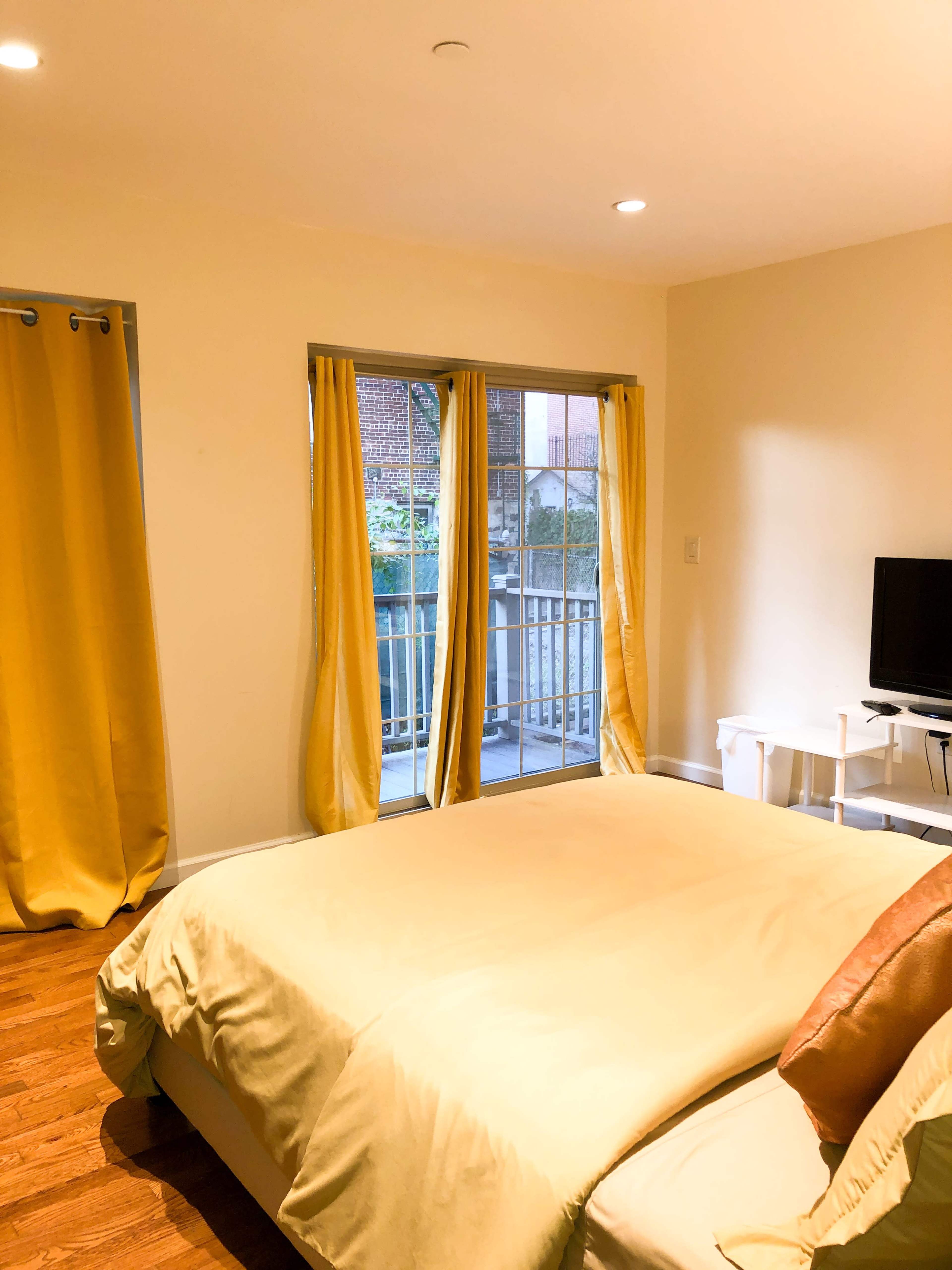 A bedroom with a bed, yellow curtains, a television on a stand, and a sliding door leading to a balcony.