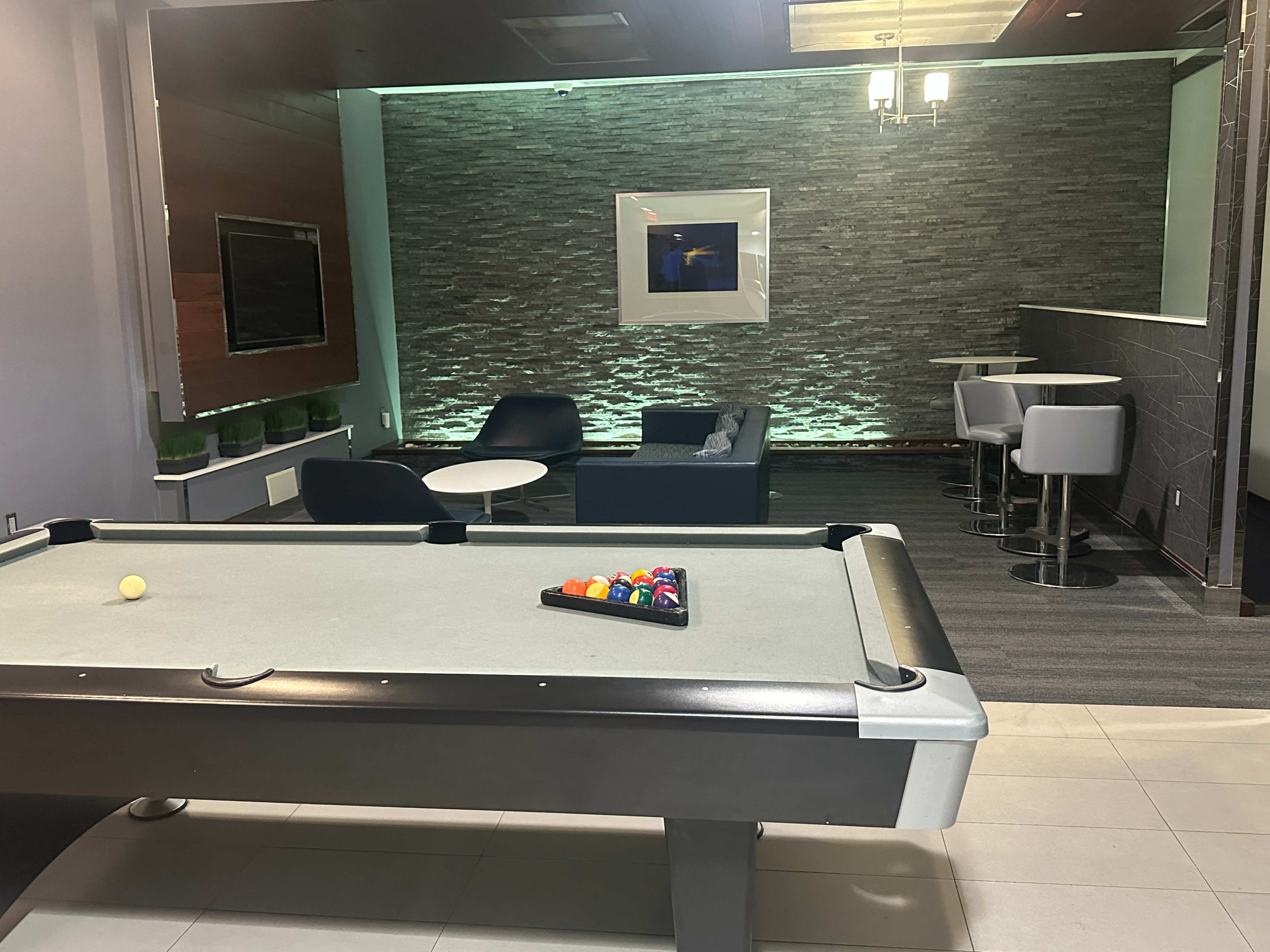 The image shows a game room with a pool table in the foreground and a seating area with modern furniture in the background.
