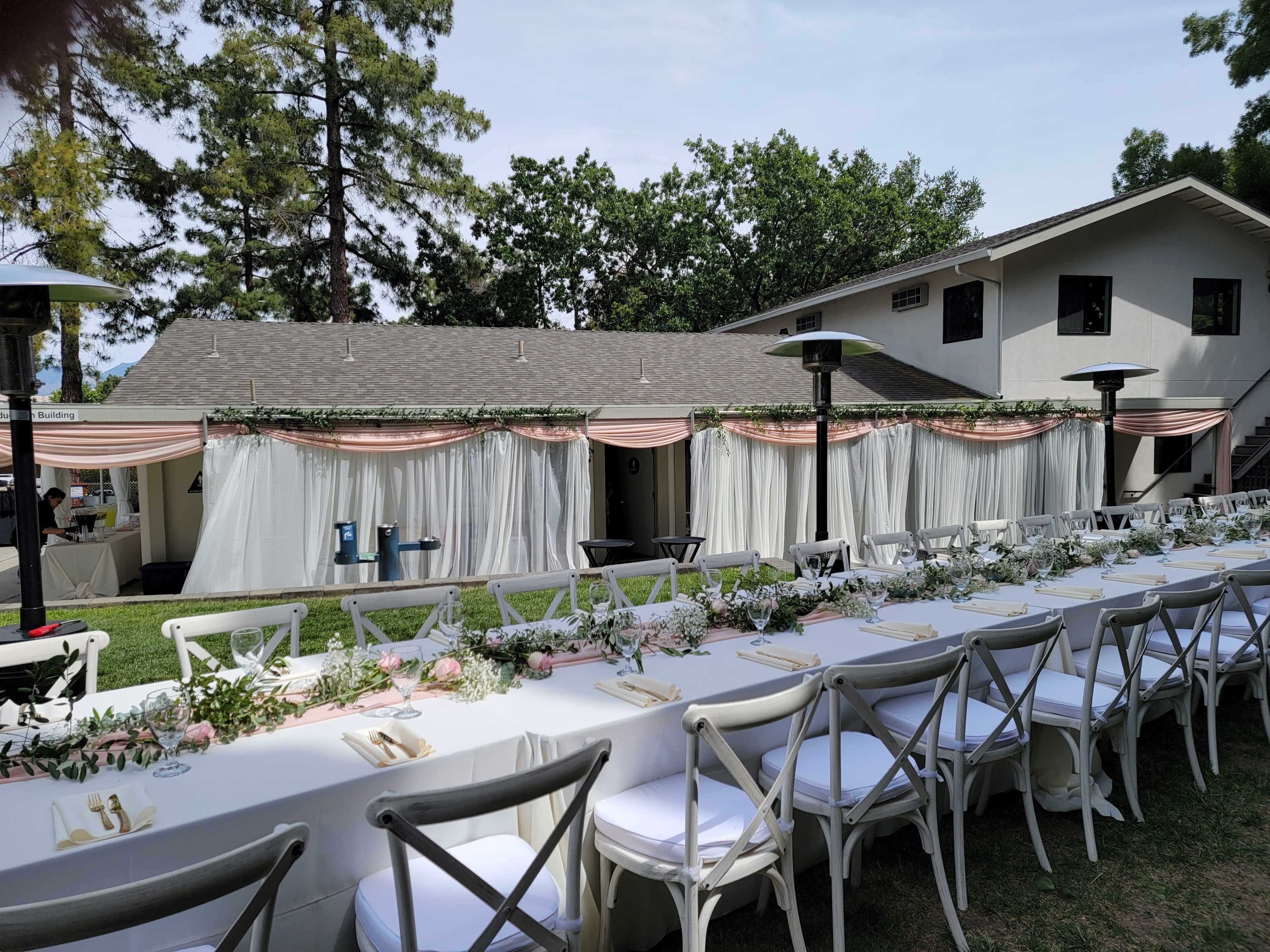 A long, elegantly set dining table is arranged outside near a building, adorned with floral decorations and draped fabric.