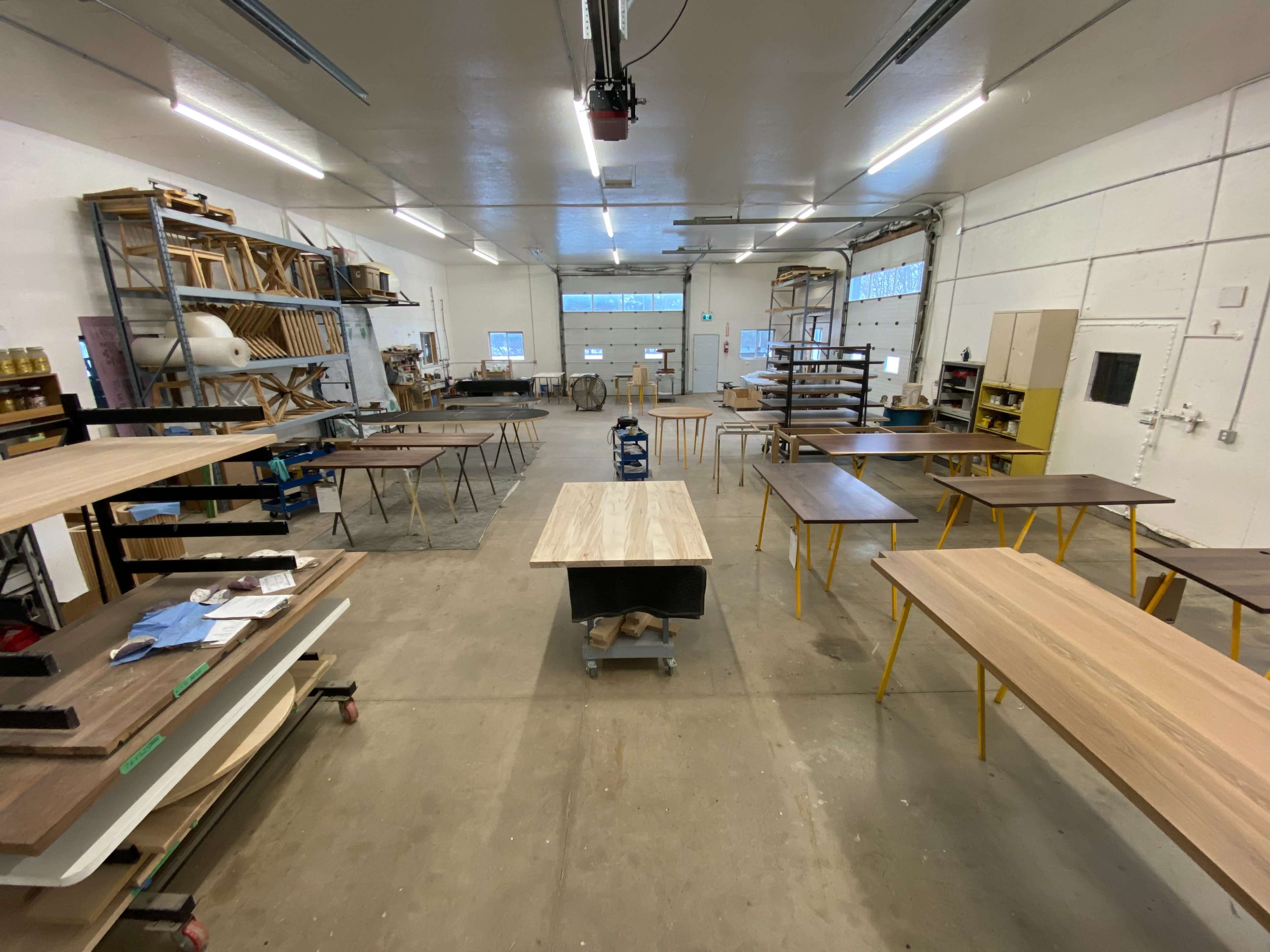 A spacious woodworking shop with several tables arranged throughout, storage shelves filled with materials, and tools positioned in the background.