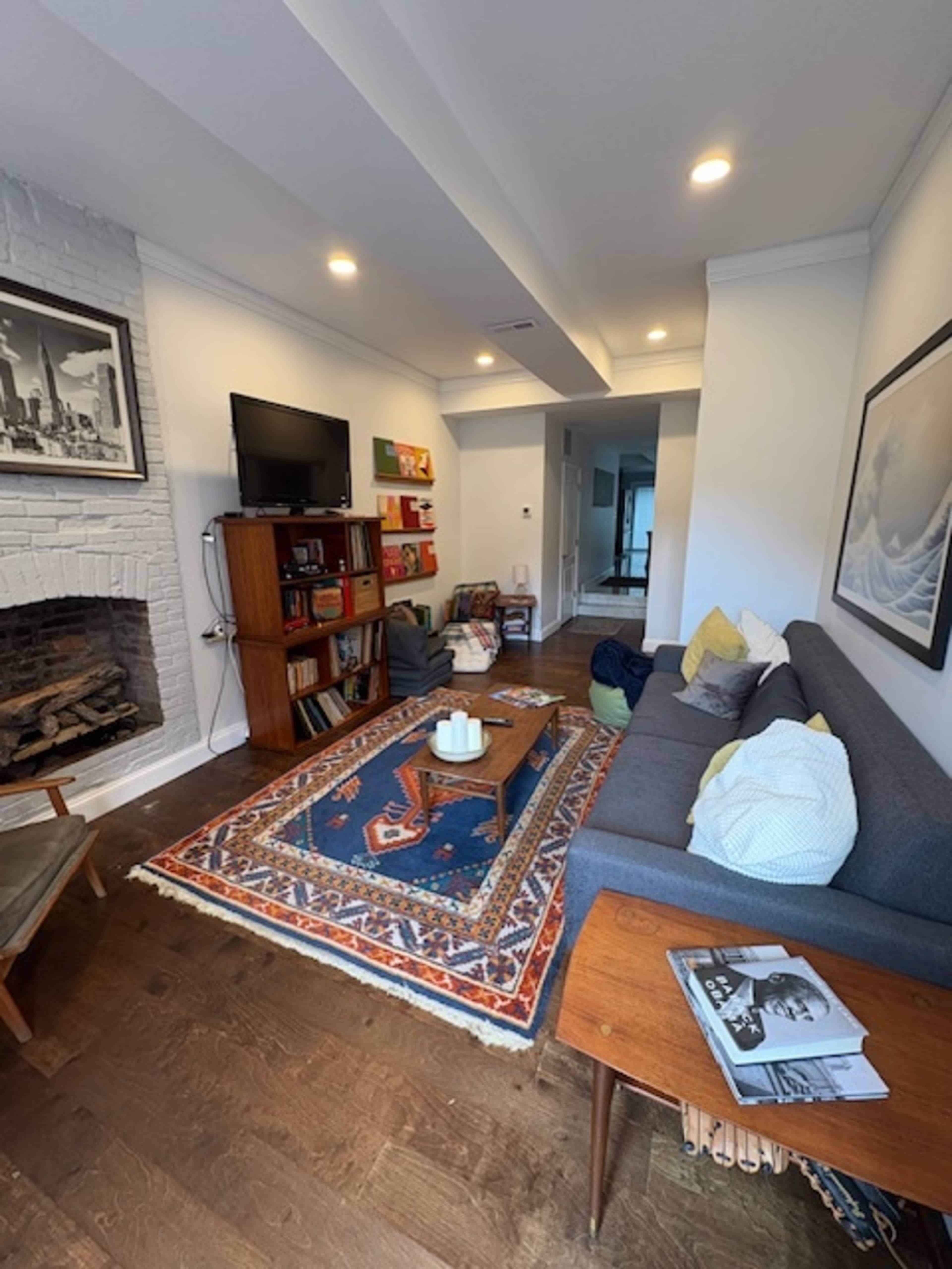 The image shows a modern living room with a gray sofa, a wooden coffee table on a colorful rug, and a bookshelf against the wall.