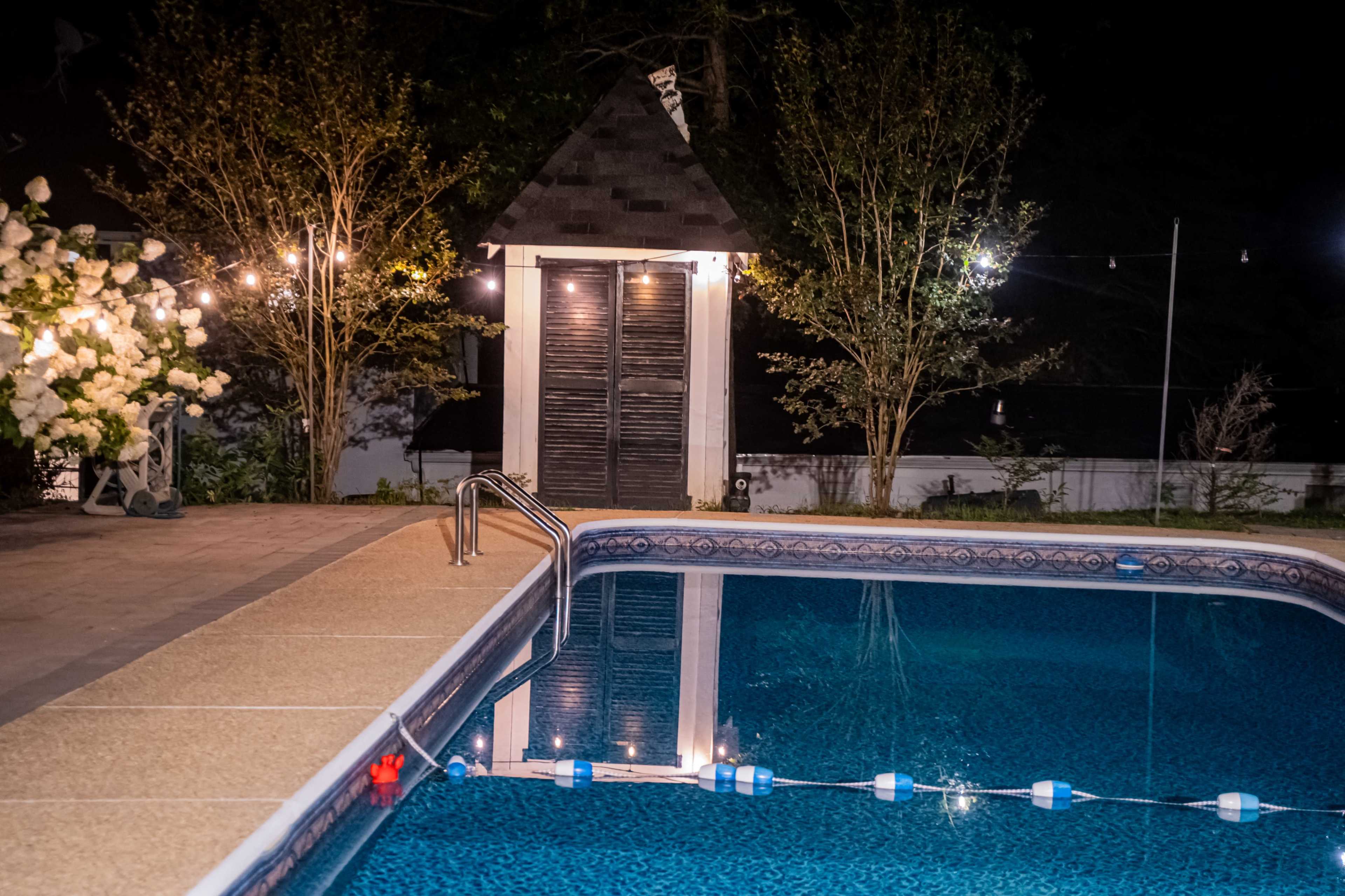 The image shows a nighttime scene of a backyard swimming pool illuminated by string lights, with a small shed and trees in the background.