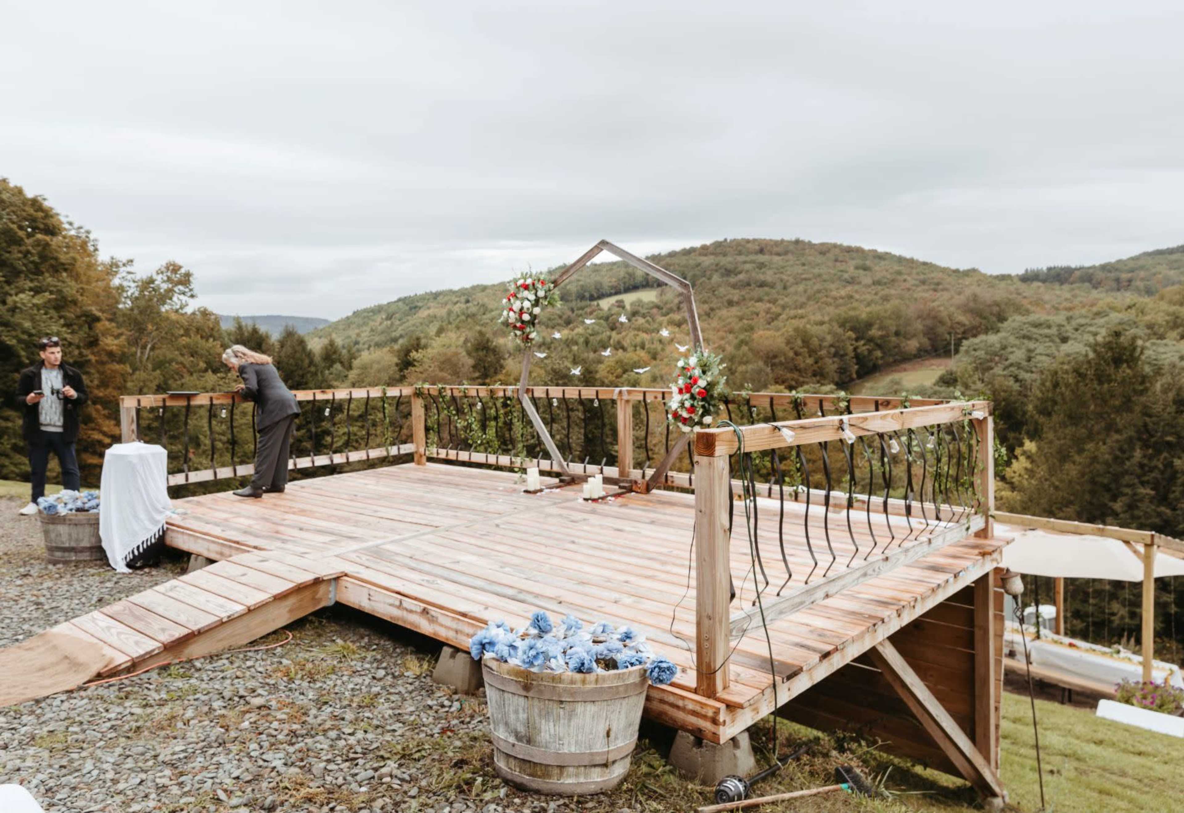 An outdoor wooden wedding altar is set up on a raised platform surrounded by greenery and mountains.