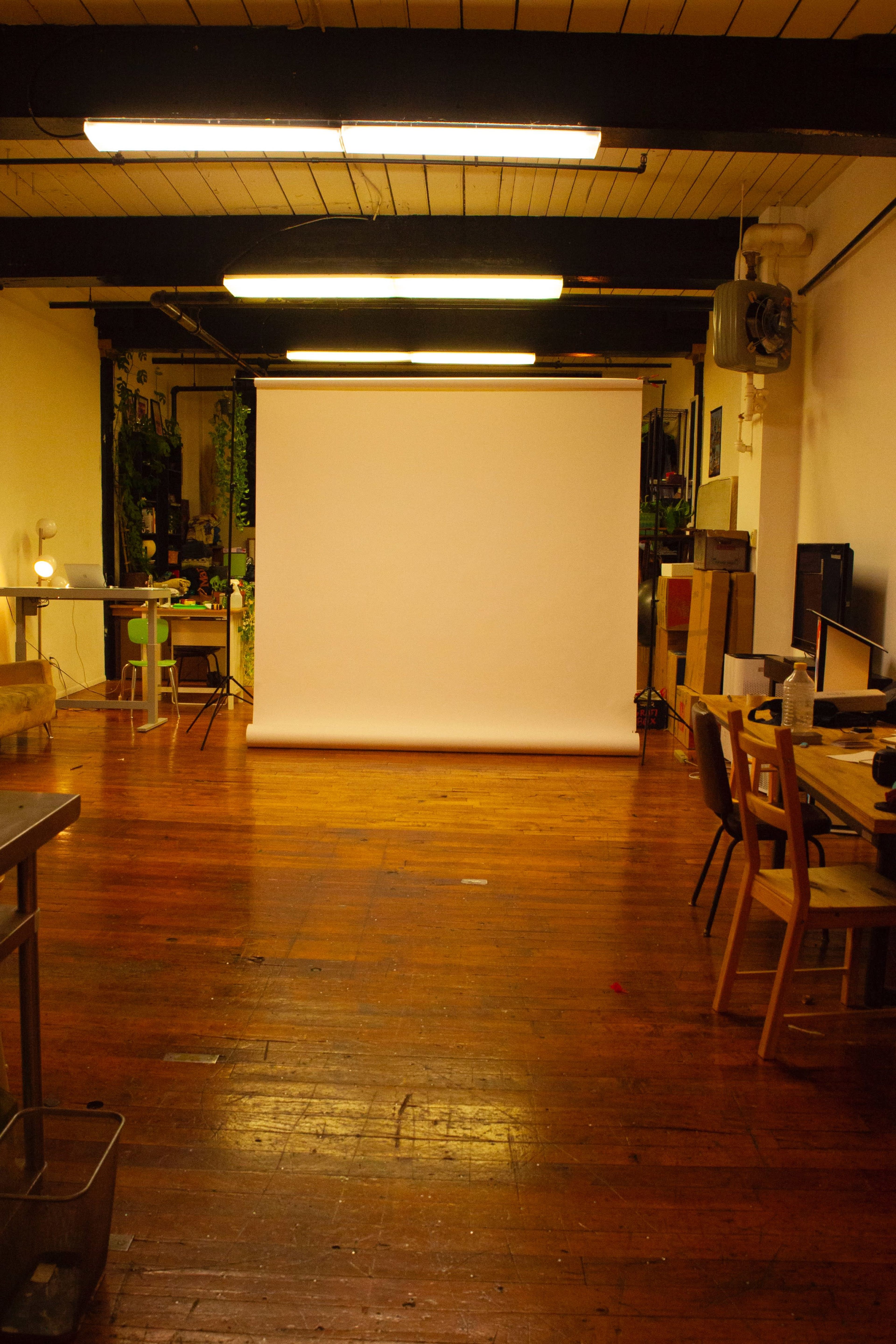 The image shows a spacious studio with wooden flooring, a blank backdrop, and various furniture pieces such as tables and chairs arranged around the room.