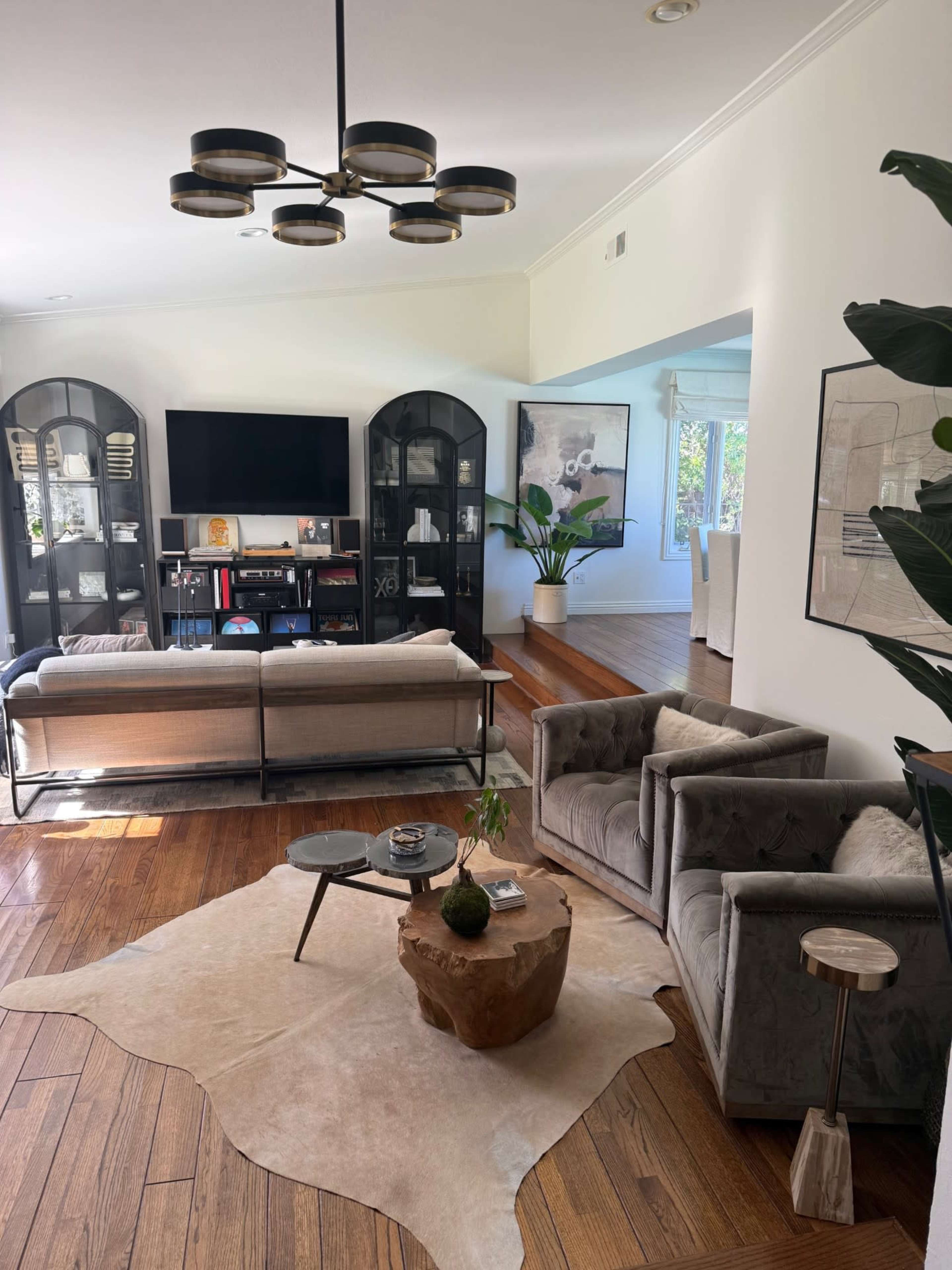 The image shows a modern living room with a large television mounted on the wall, a coffee table made of wood, and two gray armchairs positioned around a decorative area rug.