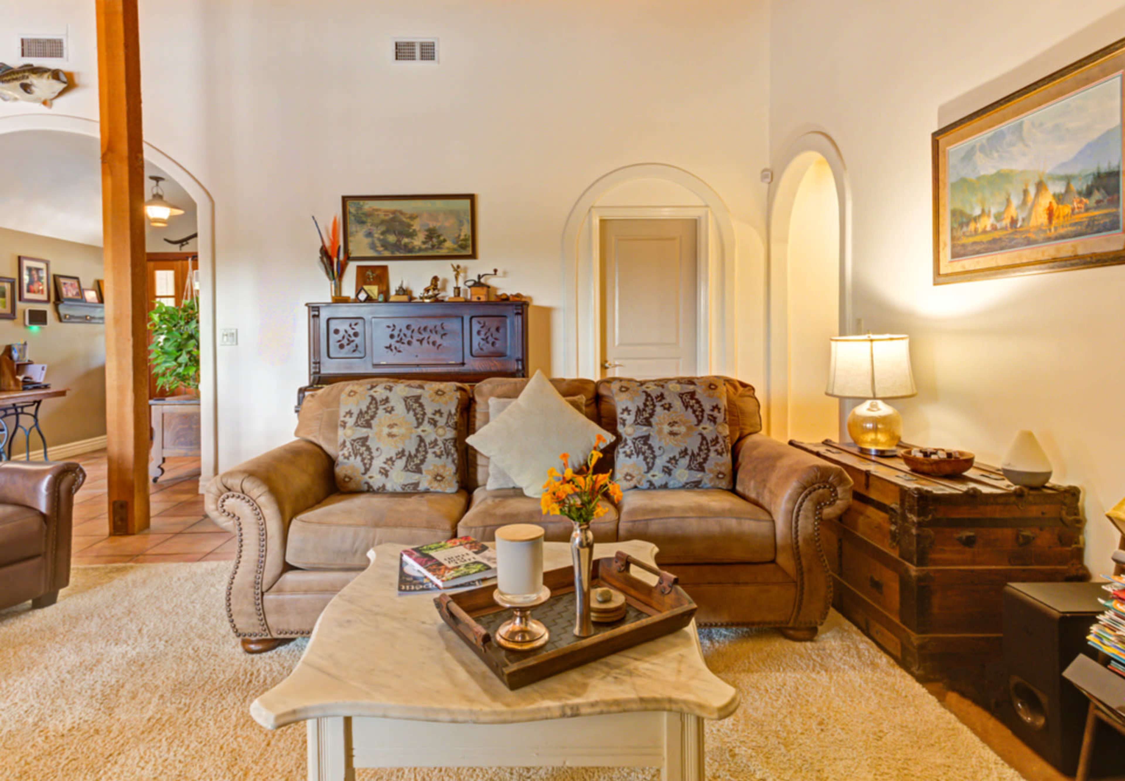 A cozy living room features a beige sofa with decorative pillows, a coffee table with a tray and books, and a lamp beside a wooden cabinet and a painting on the wall.