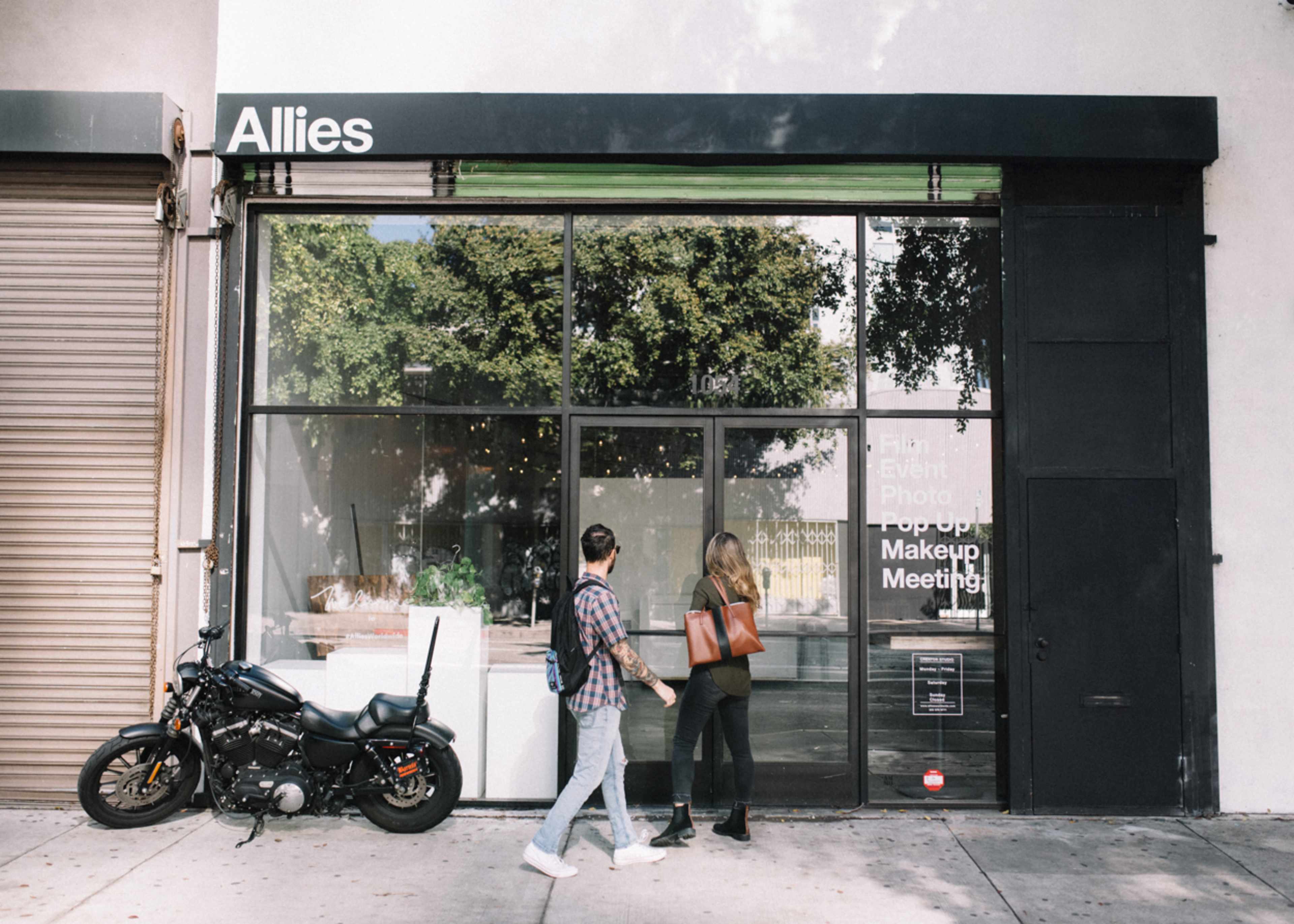 A man and a woman walk past a storefront labeled "Allies," which features a motorcycle parked outside and displays signs for events and services.