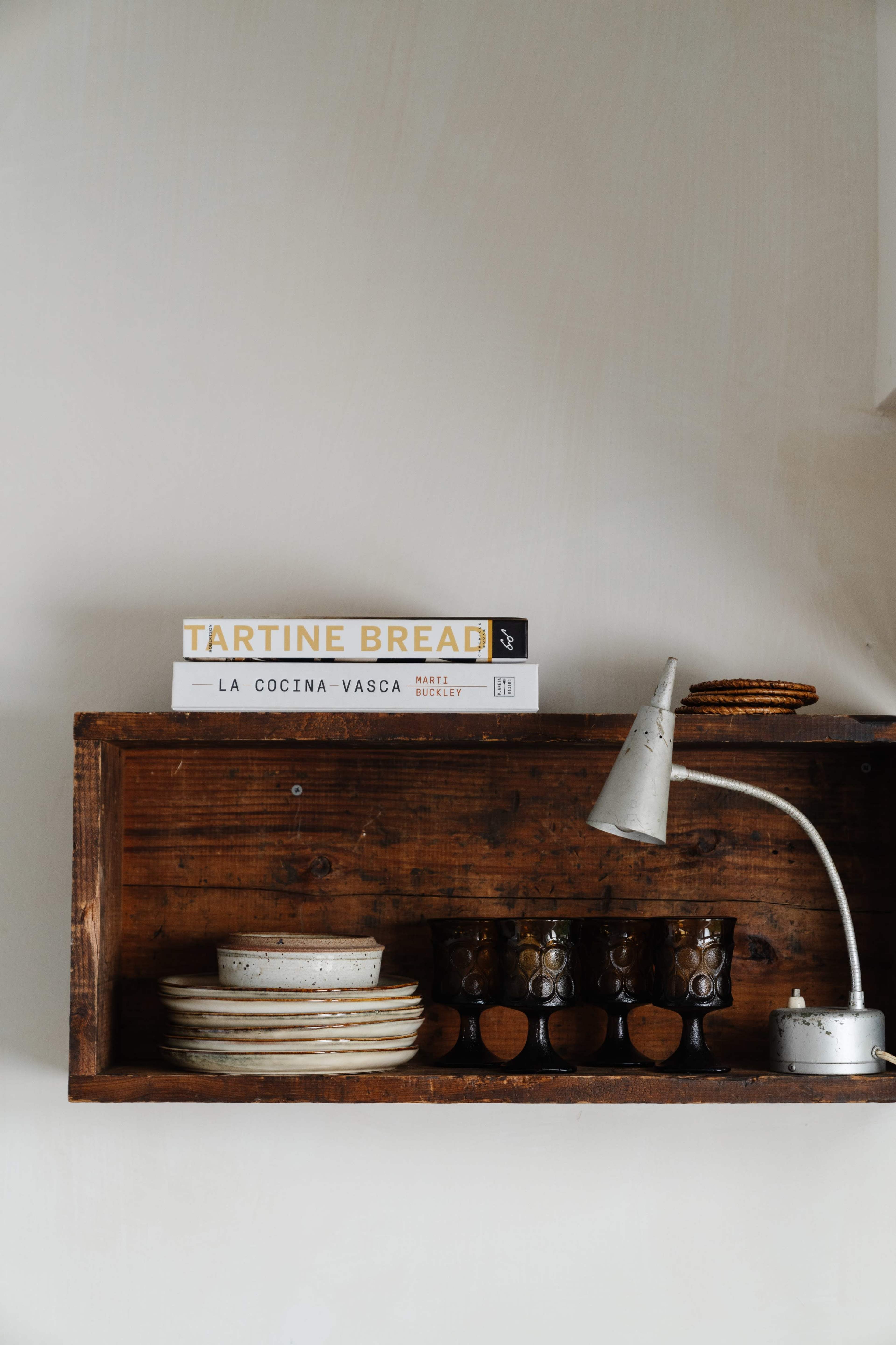 A wooden shelf displays a stack of cookbooks, ceramic plates, and three dark glass cups under a small lamp.