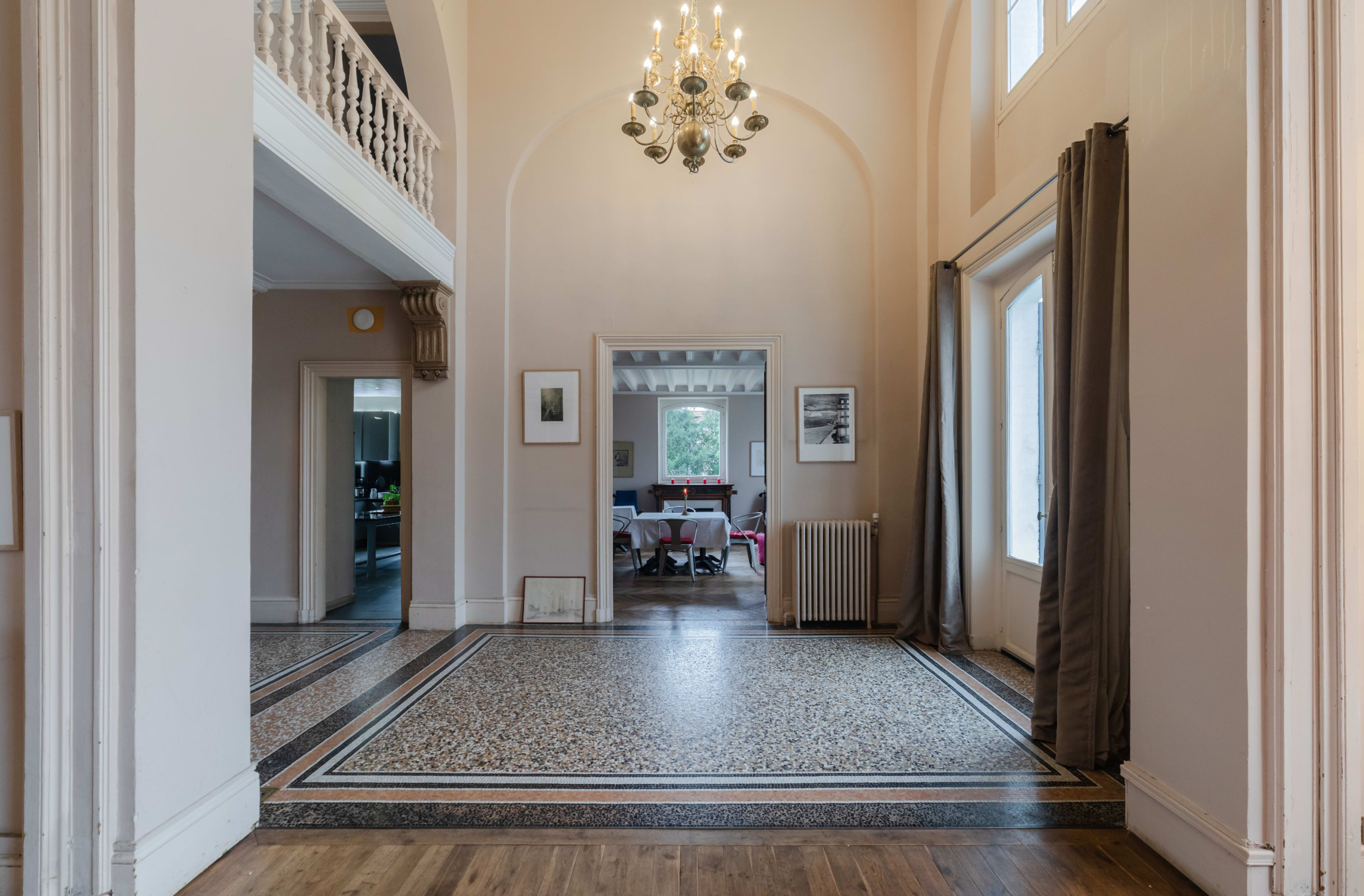 A spacious entryway with patterned tile flooring and tall windows, leading to a room illuminated by a chandelier.