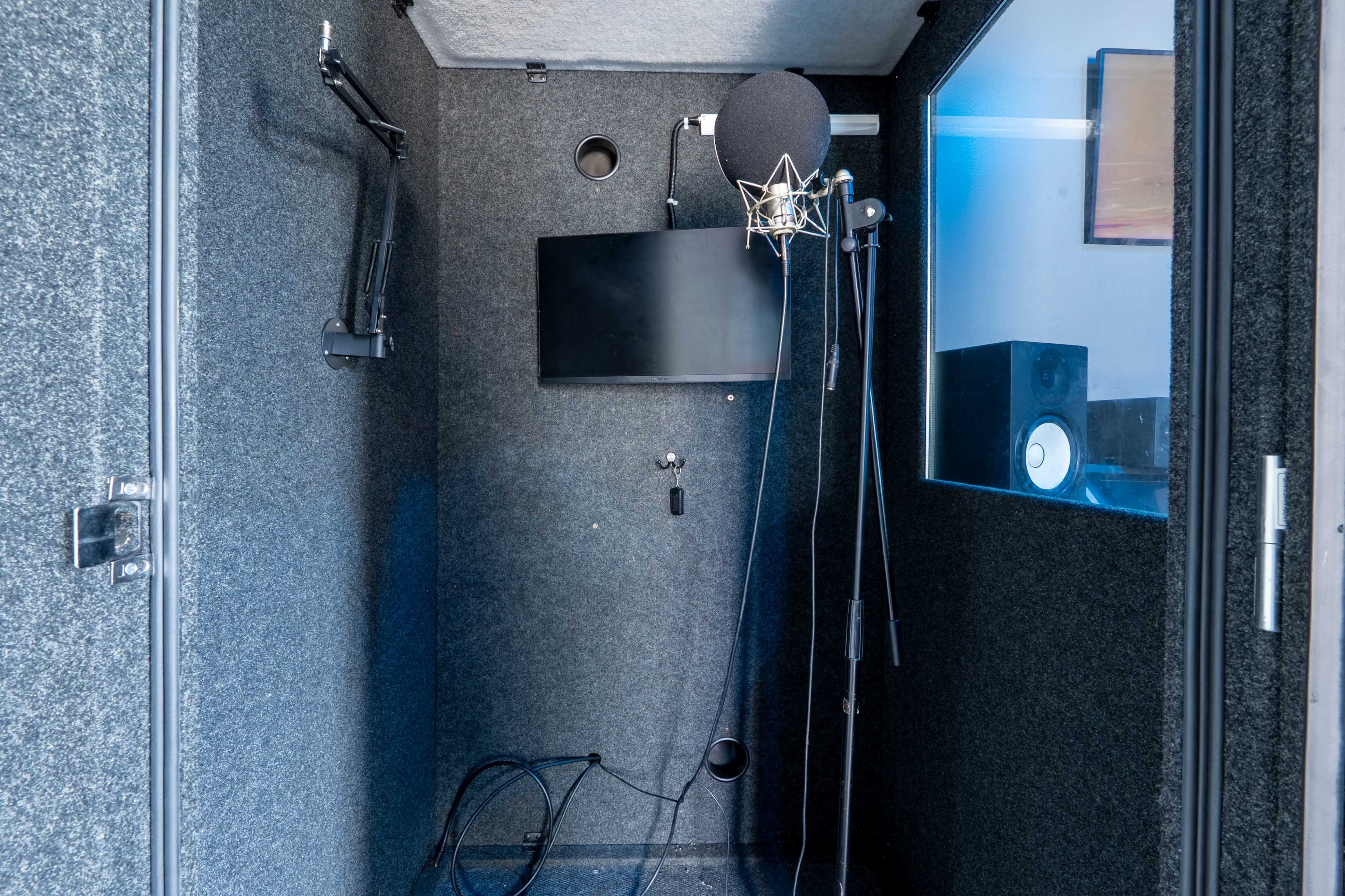 The image shows a soundproof recording booth with a microphone, a mounted monitor, and audio equipment inside.