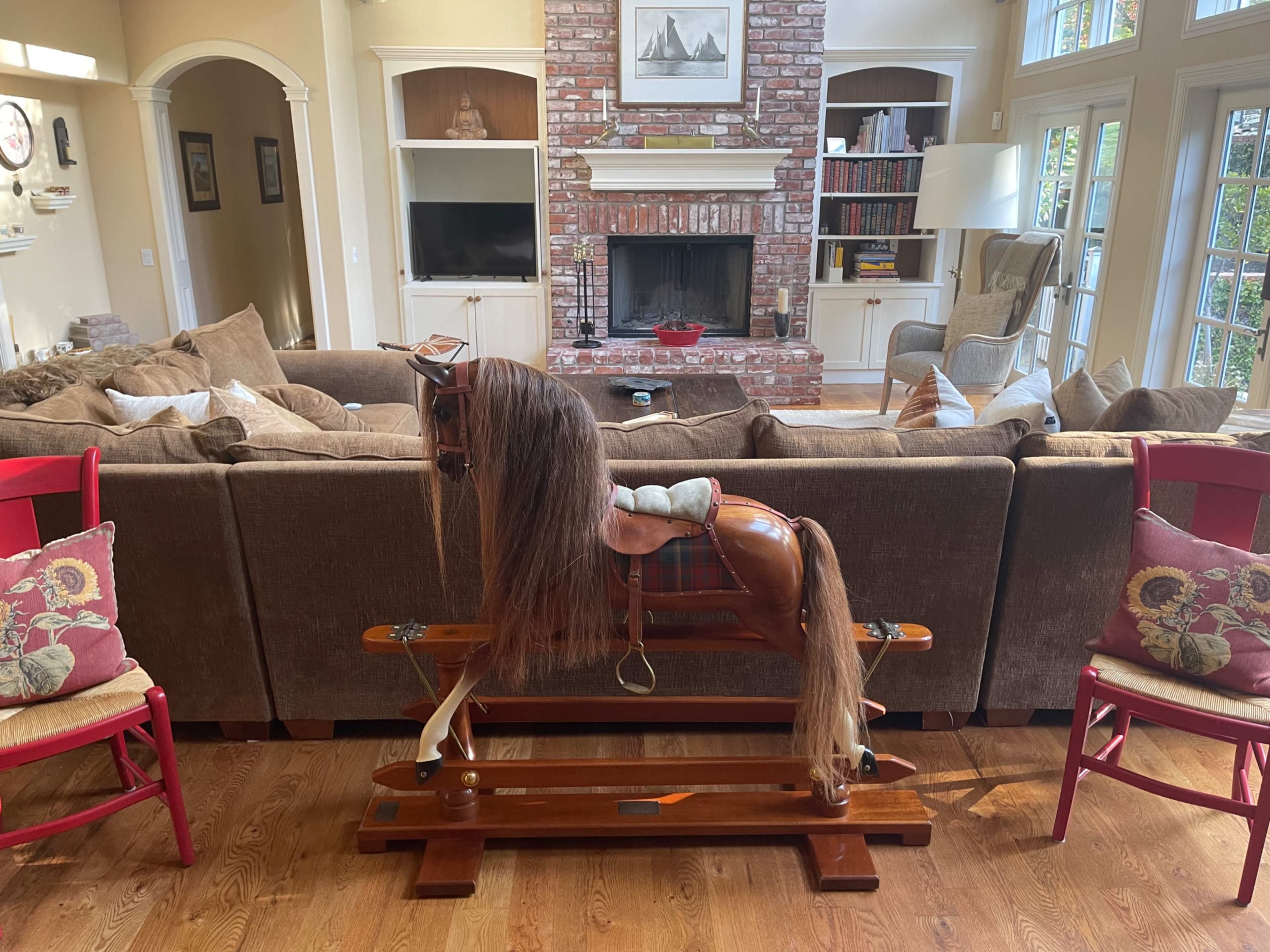A vintage rocking horse is positioned in front of a large brown sofa in a living room with a stone fireplace and built-in shelving.