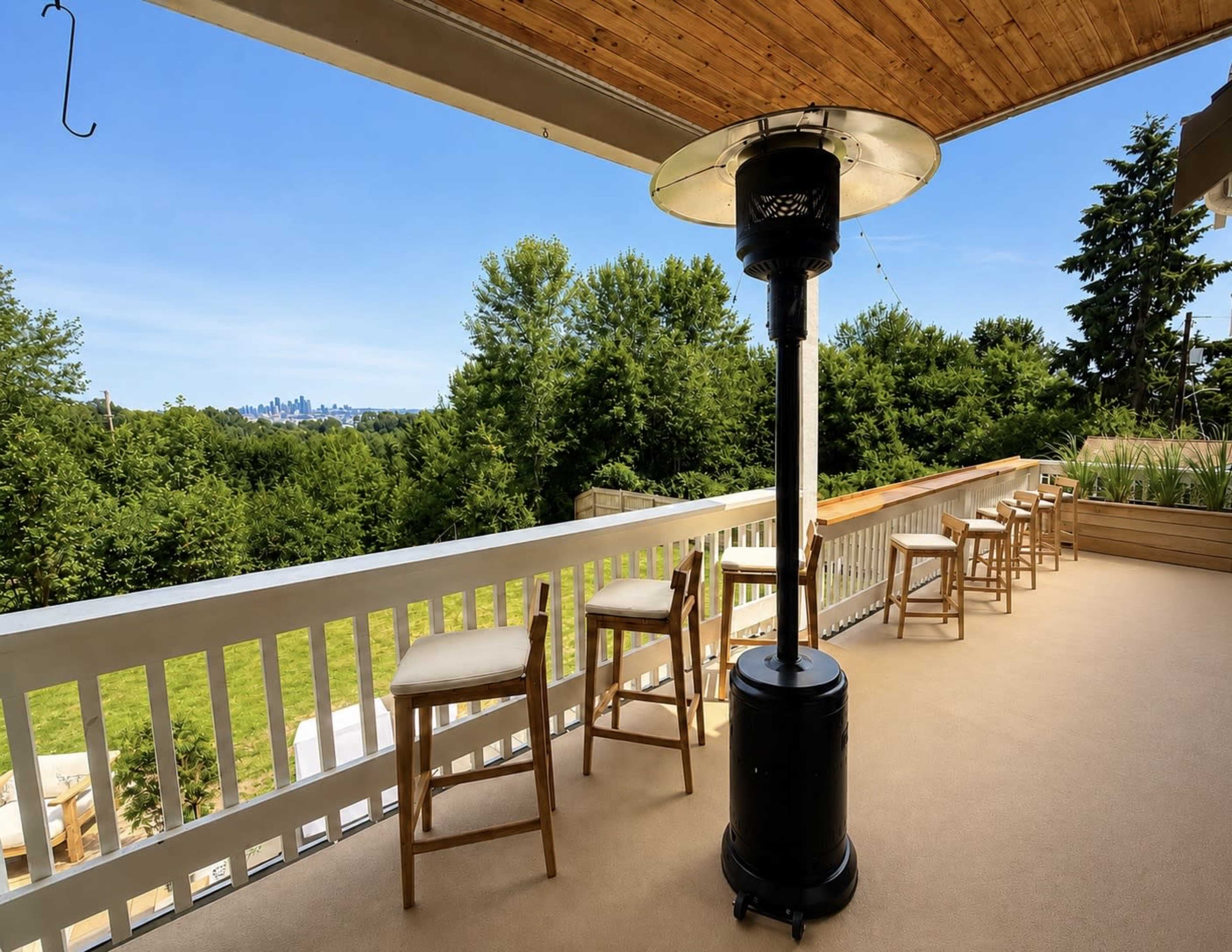 The image shows a balcony with several bar stools and a patio heater overlooking a green landscape and a city skyline in the distance.