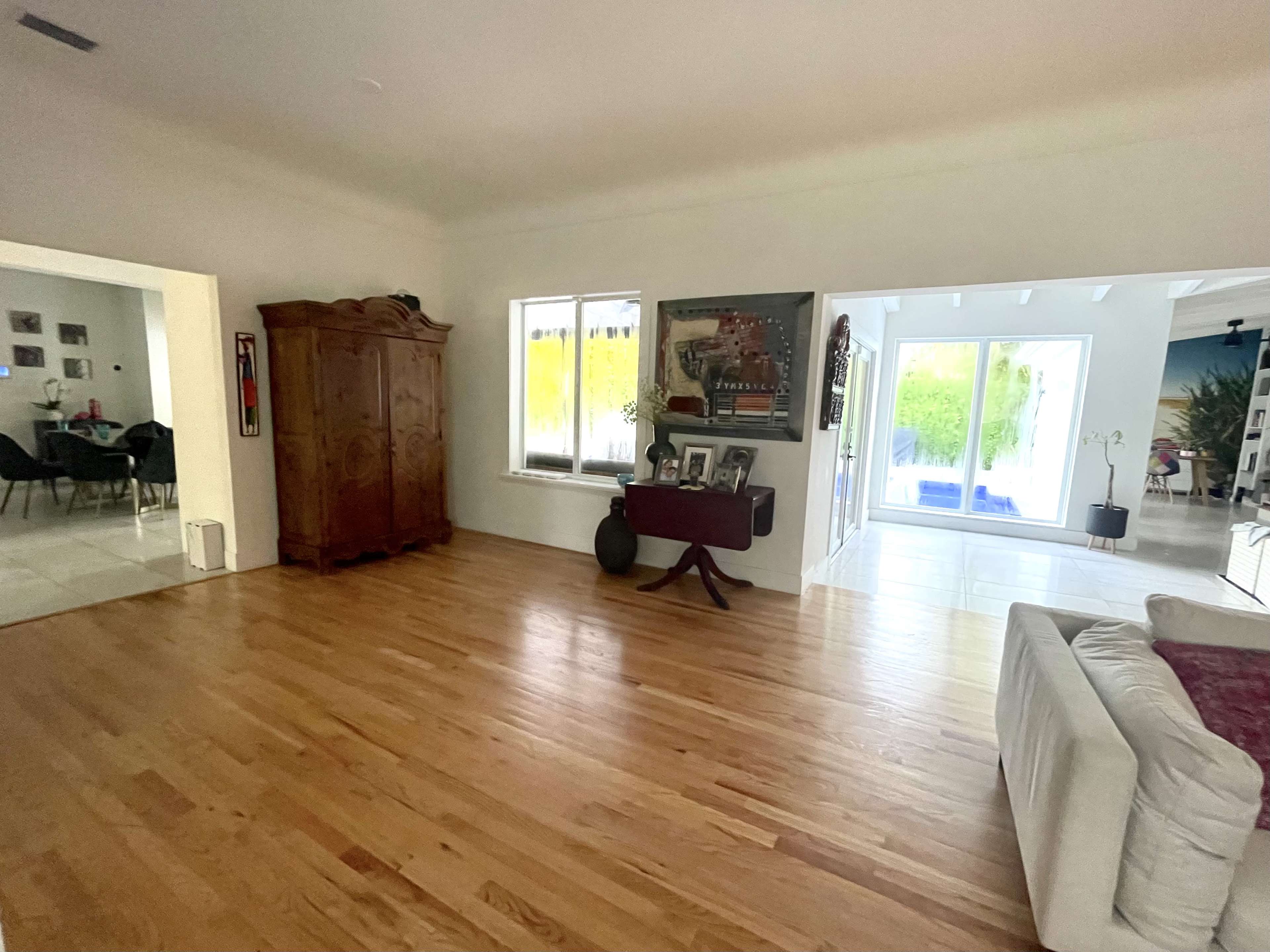 A spacious living room with hardwood floors, large windows, and a mix of furniture including a wooden cabinet and a white couch.