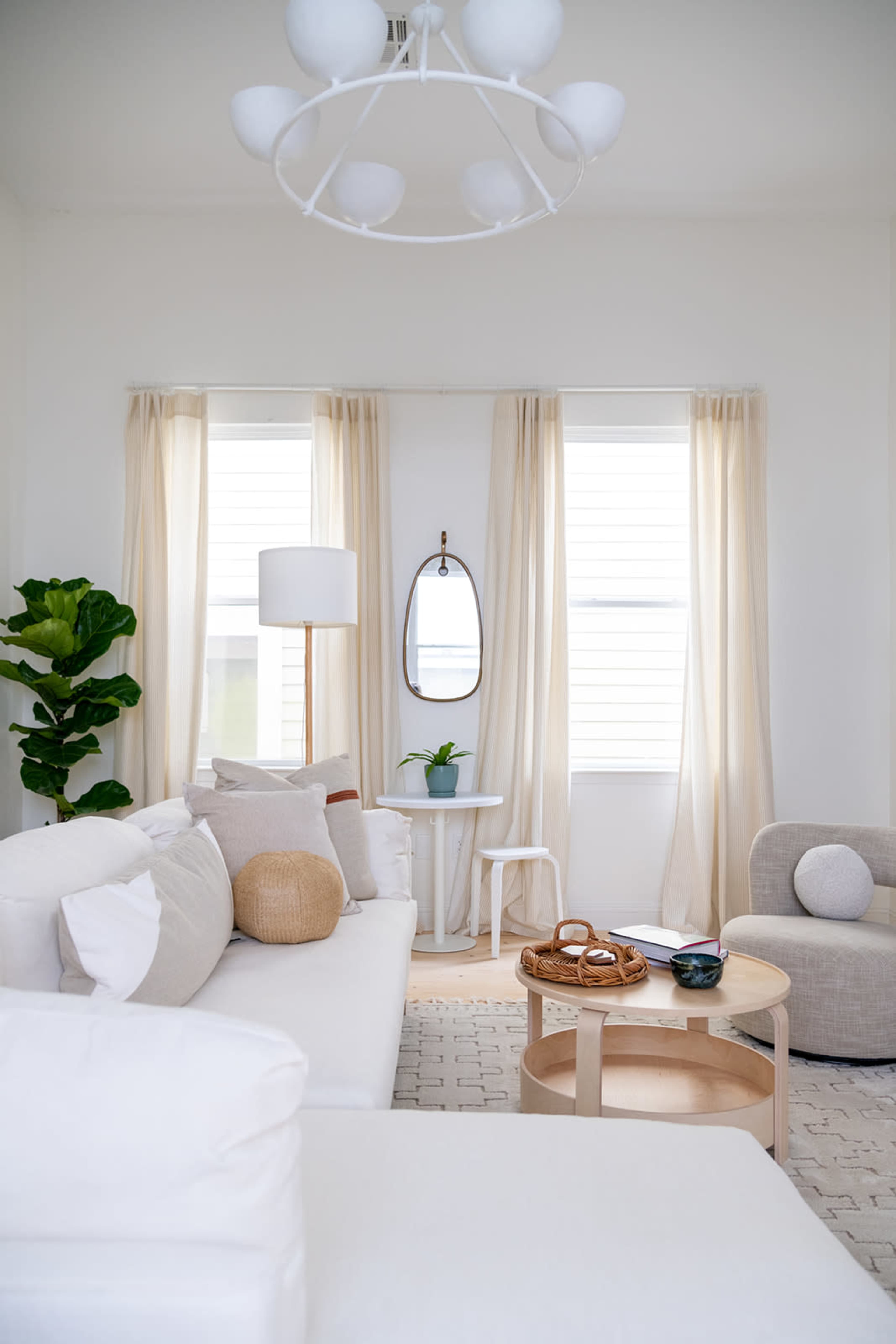 A bright living room features a white sofa, a circular wooden coffee table, a lamp, and a mirror opposite two large windows with sheer curtains.