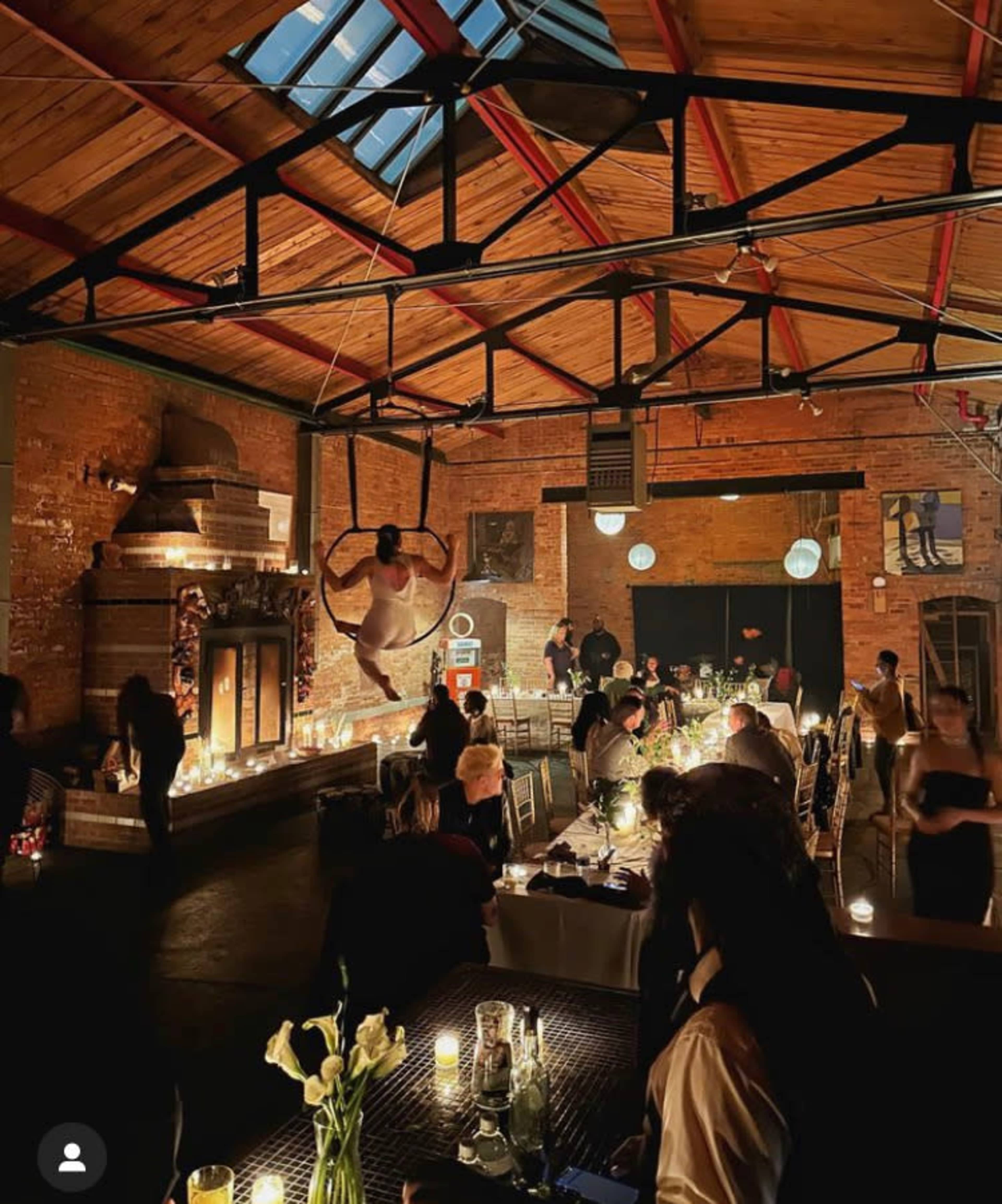 An indoor restaurant with a performer suspended from the ceiling, surrounded by diners seated at tables illuminated by candlelight.