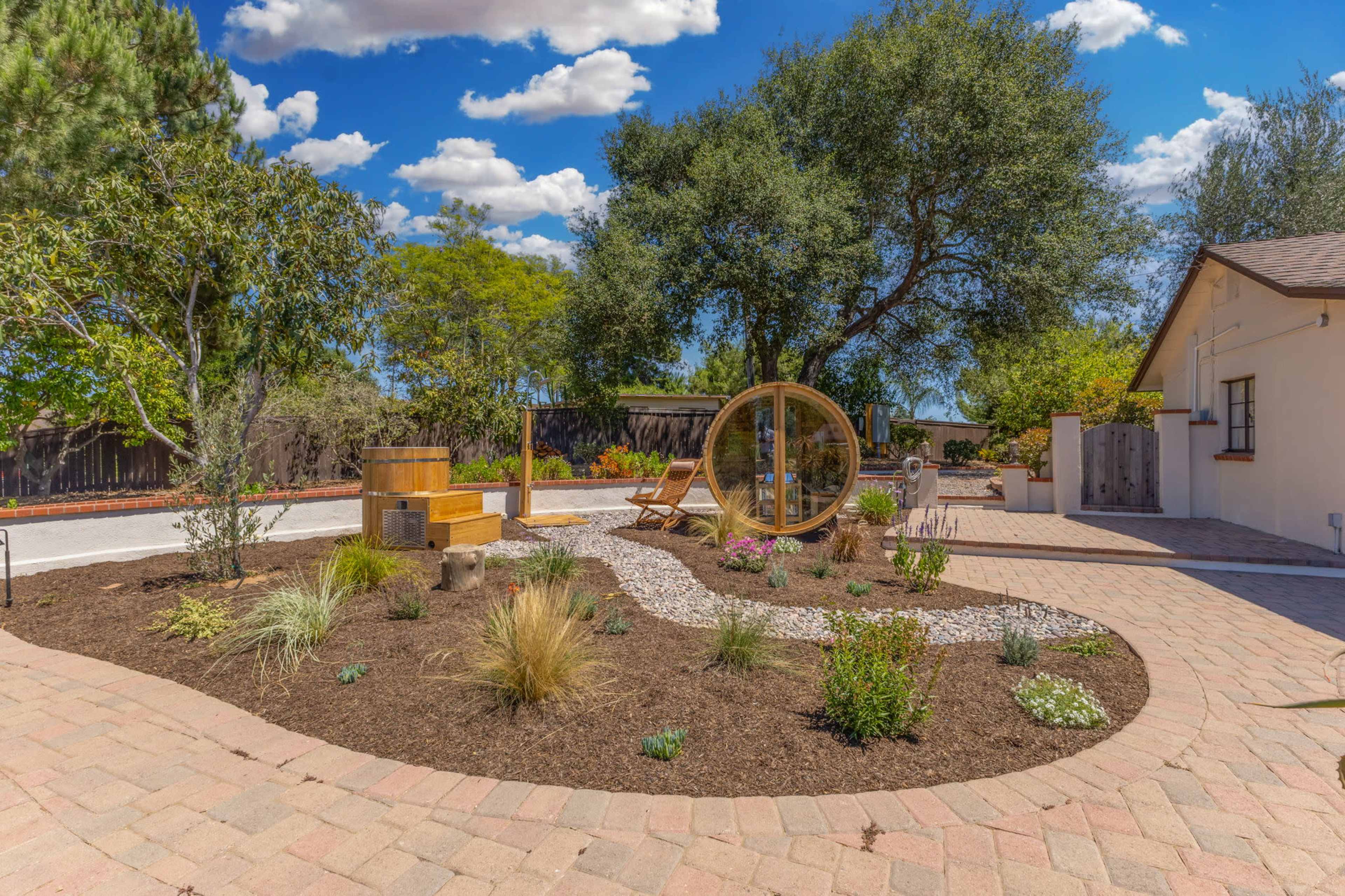 A landscaped outdoor space features a circular wooden structure, two lounge chairs, and various plants surrounded by a stone pathway.