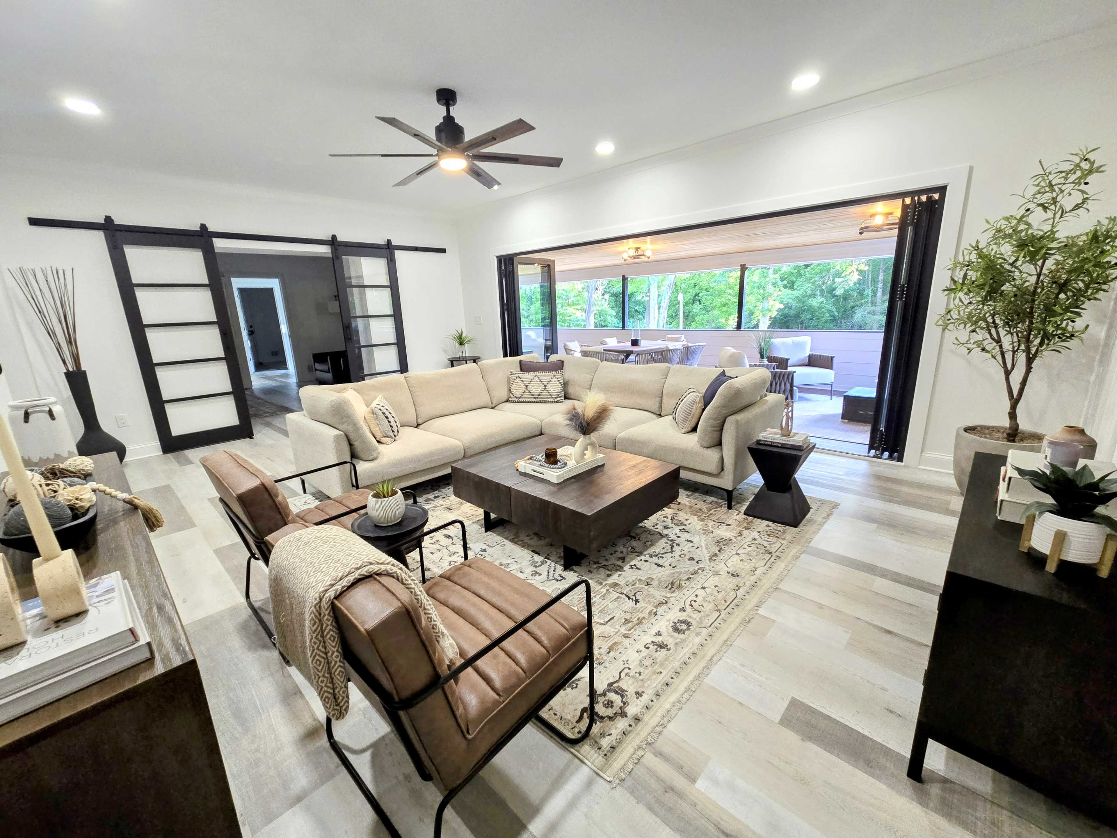 The image shows a modern living room with a large beige sectional sofa, a coffee table, and two brown chairs, all arranged near a sliding door that opens to a patio area.