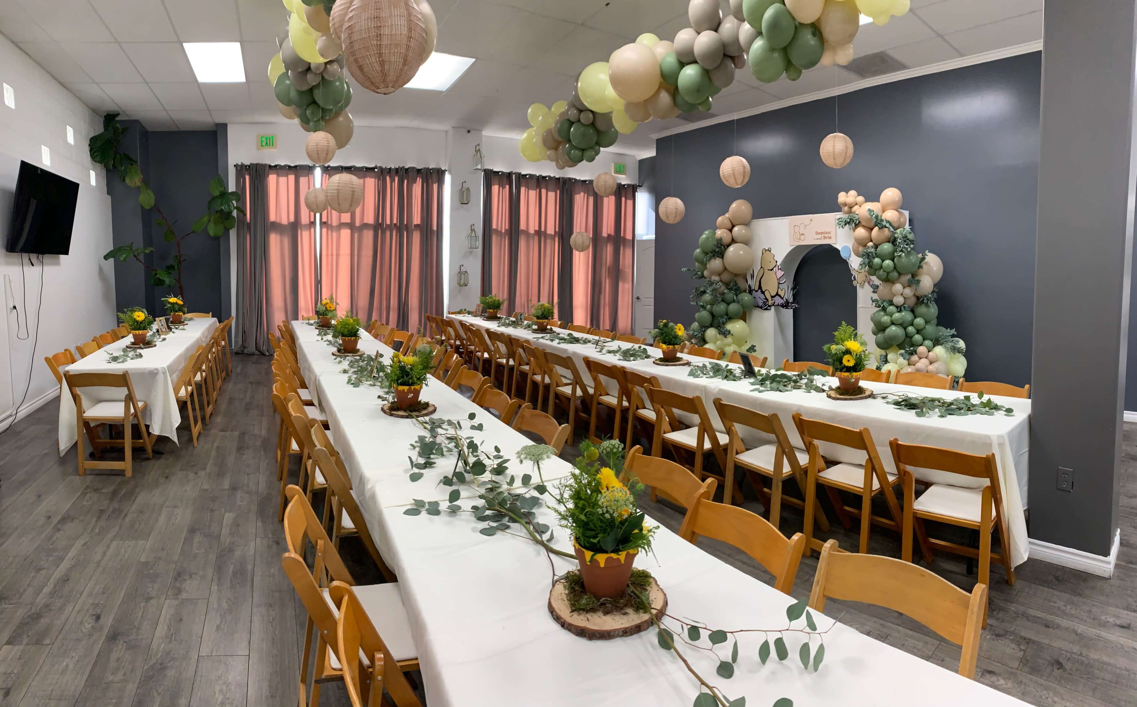 A spacious event hall features long tables adorned with floral centerpieces and greenery, surrounded by balloon decorations and pendant lights.