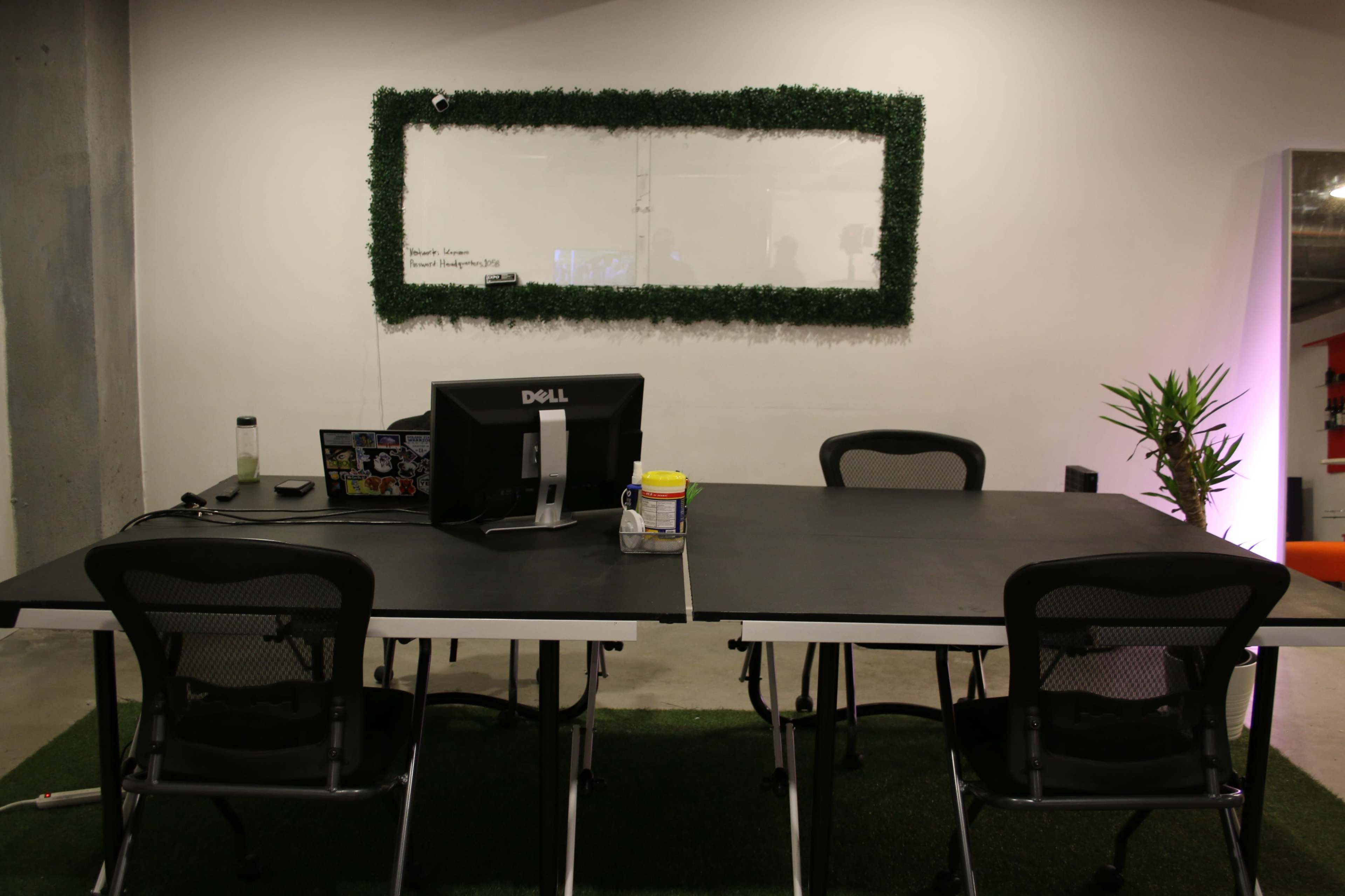A modern office space features a large table with chairs, a computer monitor, and a wall adorned with a green decorative frame.