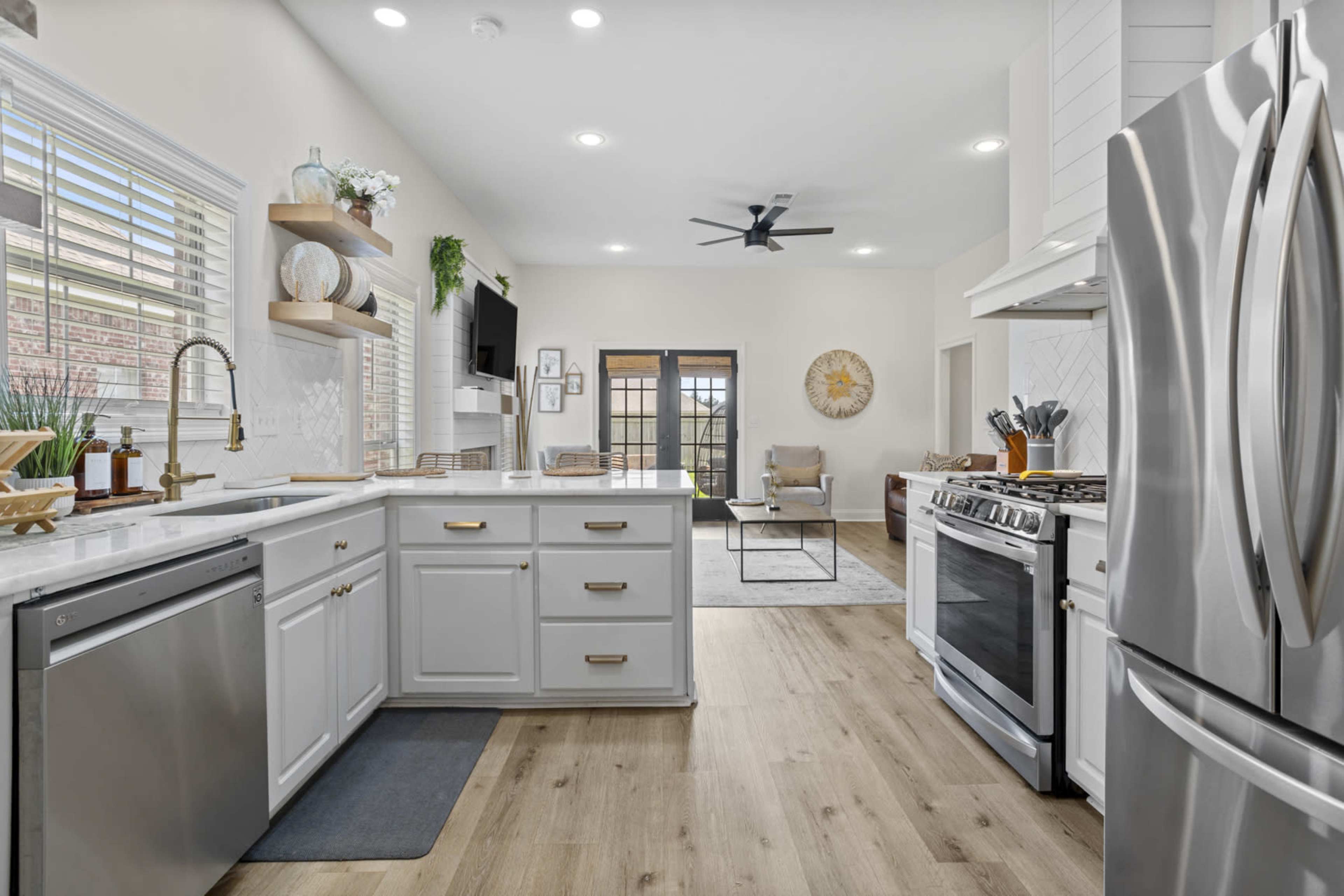 The image shows a modern kitchen with white cabinetry, stainless steel appliances, and a light wood floor, leading into a living area with a chair and a decorative sun wall piece.