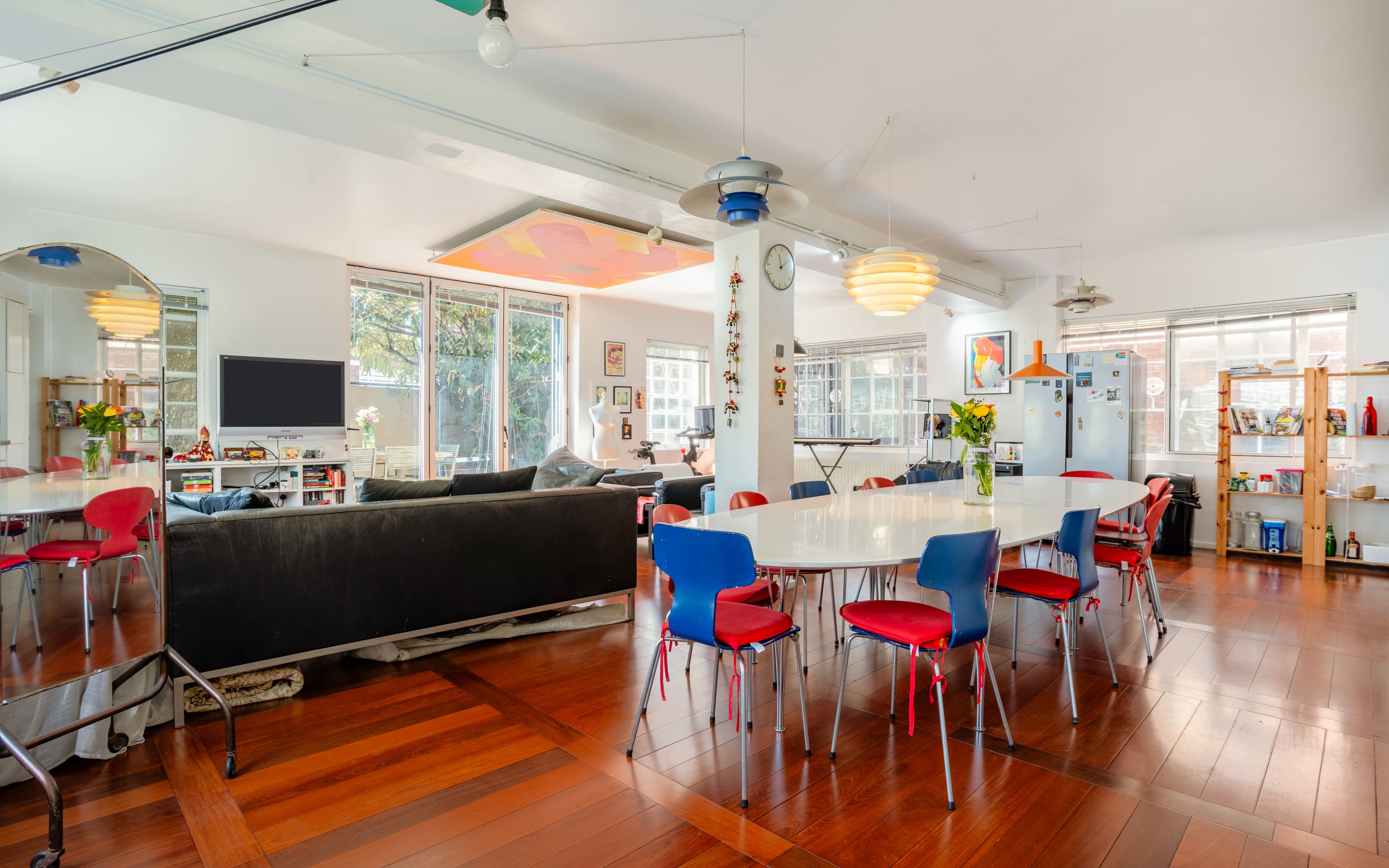 The image shows an open-concept living and dining area featuring a large white table surrounded by colorful chairs, large windows, and various decorative elements.