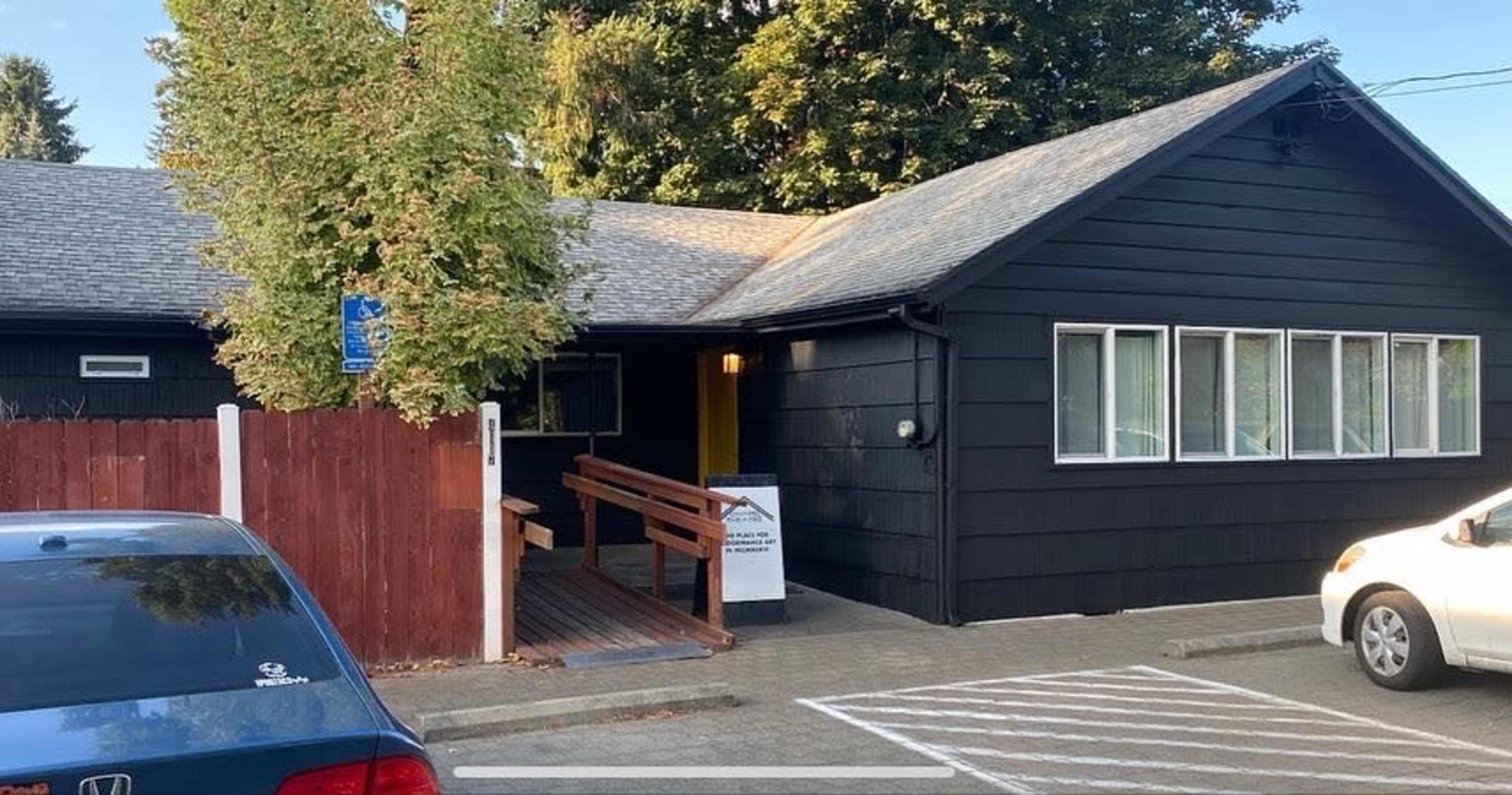 A black wooden building with multiple windows and a ramp leads to the entrance, surrounded by trees and parked cars.