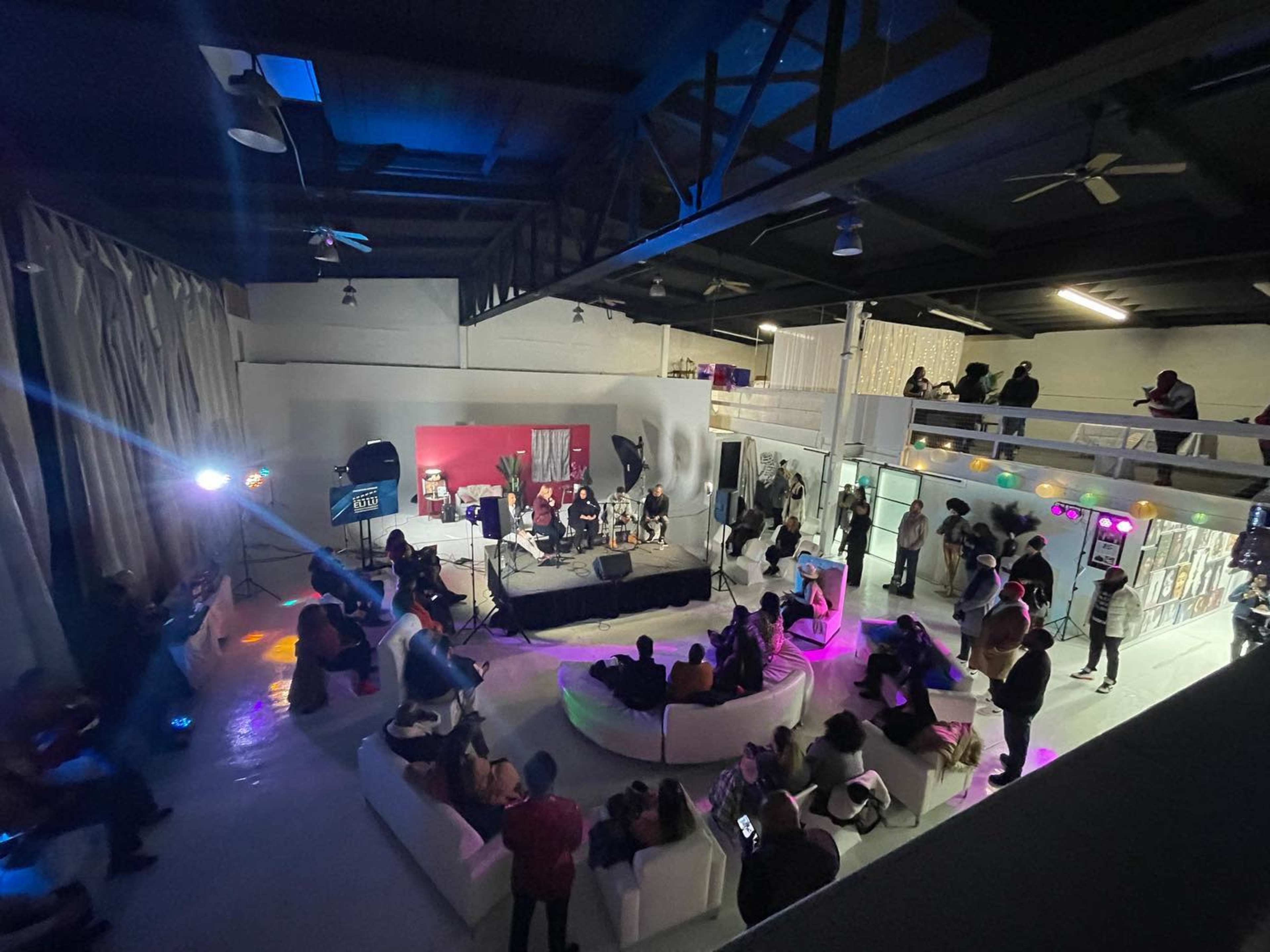 A diverse audience watches a live performance in a spacious, modern venue with elevated seating and various light setups.