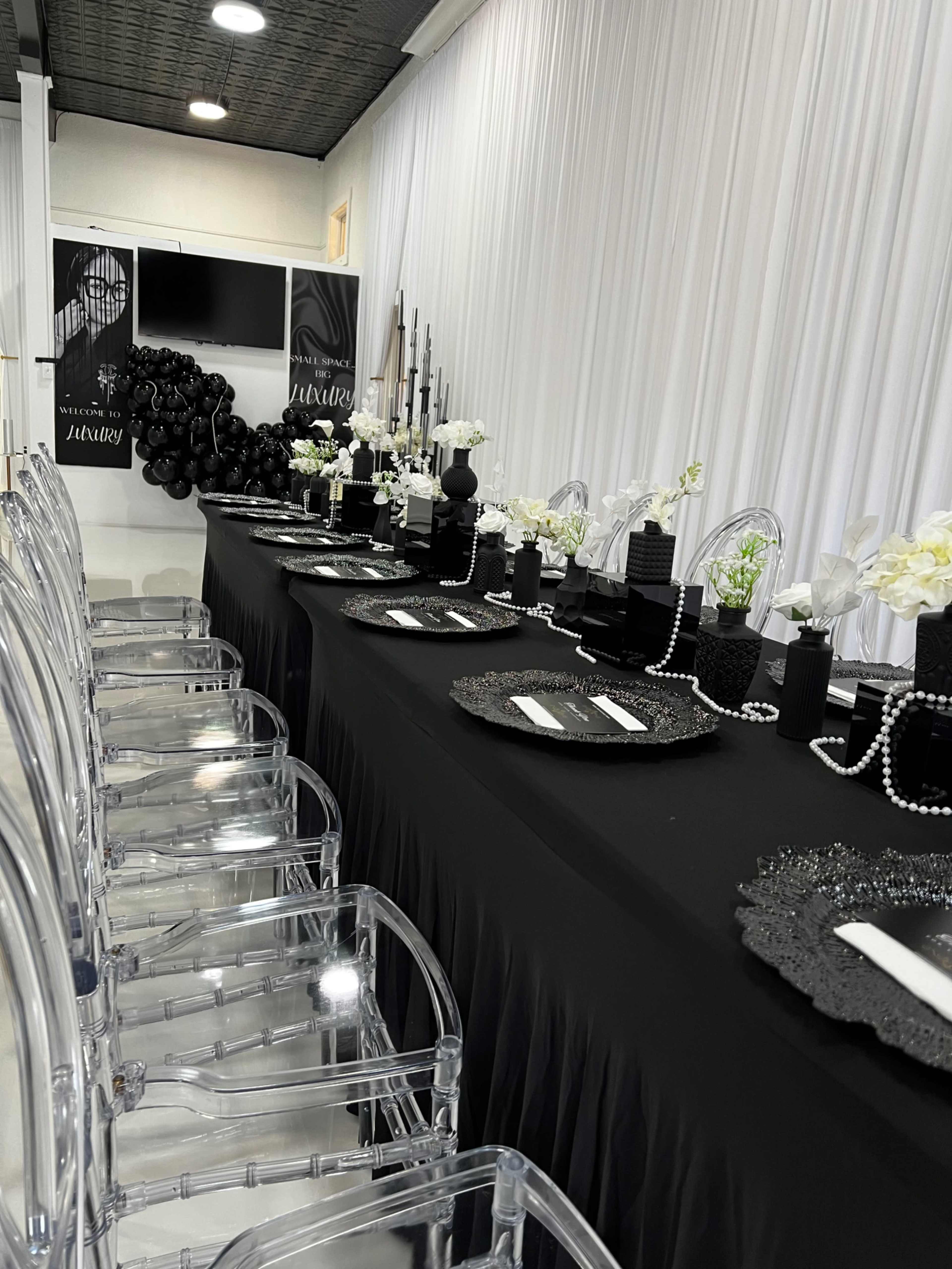 A long dining table is elegantly set with black tablecloth, clear chairs, and centerpieces of flowers and decorative items against a backdrop of white drapery.