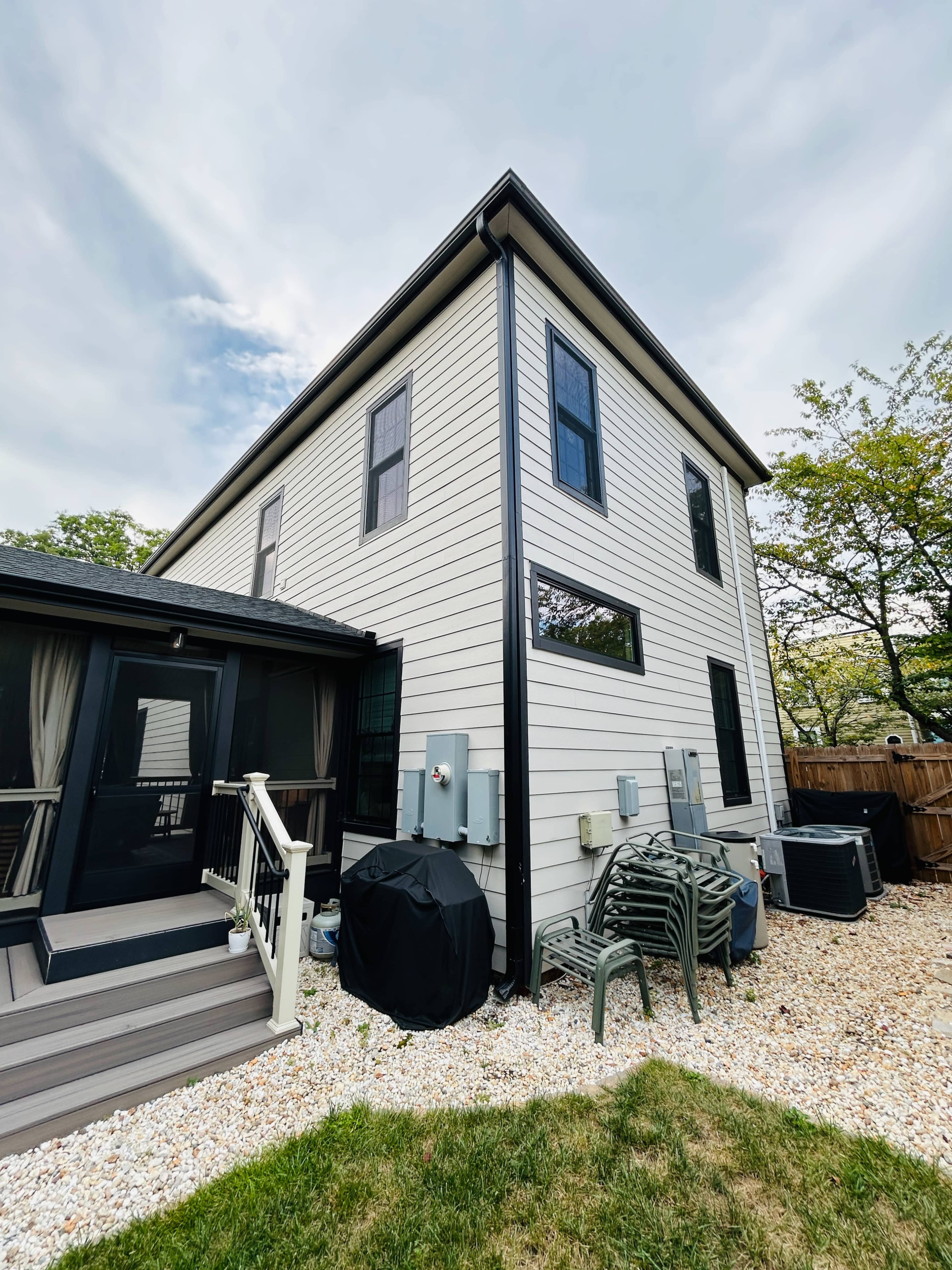 The image shows the exterior corner of a two-story house with a gray siding, a deck with steps, outdoor appliances, and a landscaped yard.