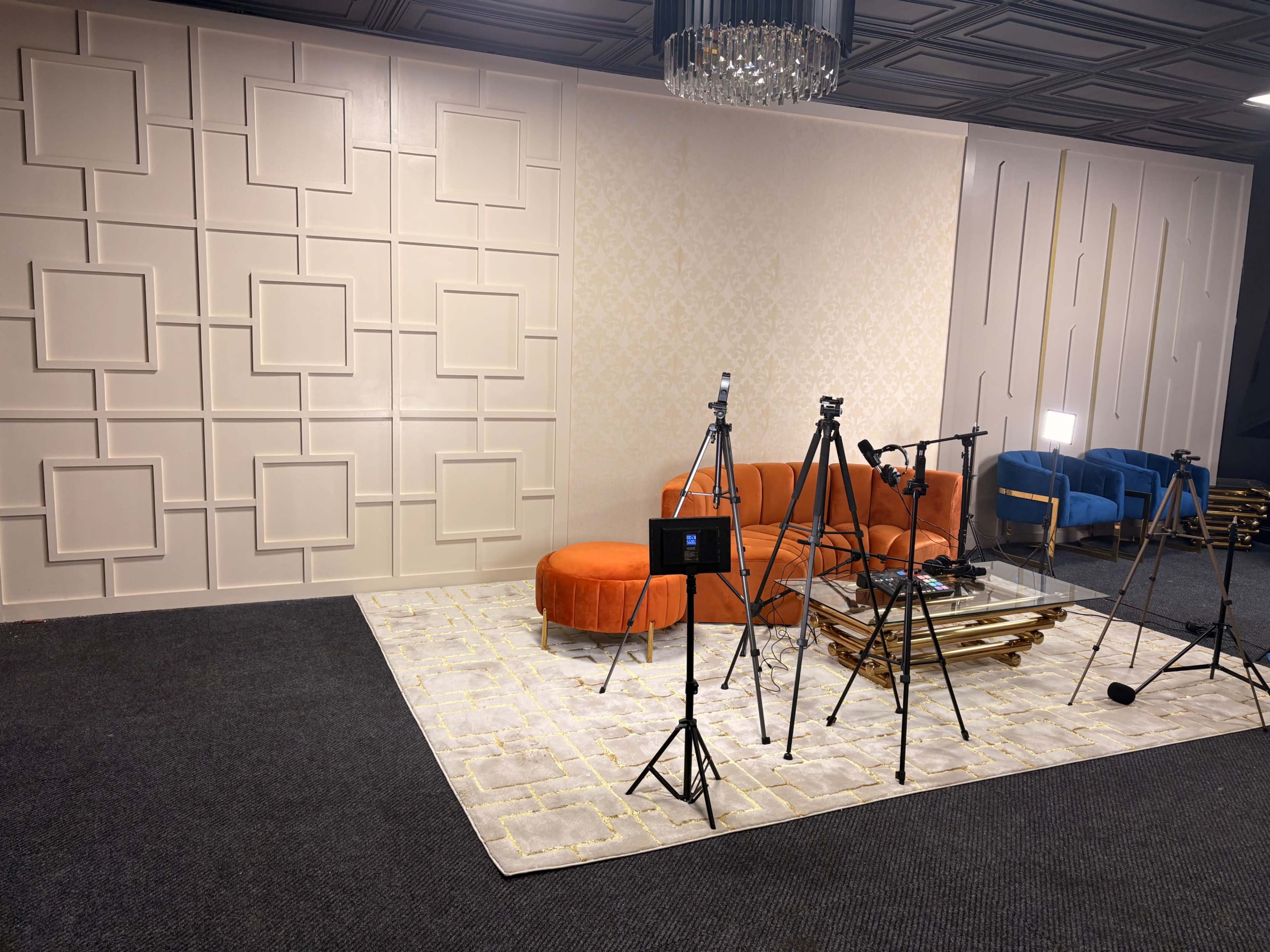 The image shows a styled indoor studio setting with an orange circular sofa, a blue armchair, a glass coffee table, and several camera tripods positioned on a patterned rug.