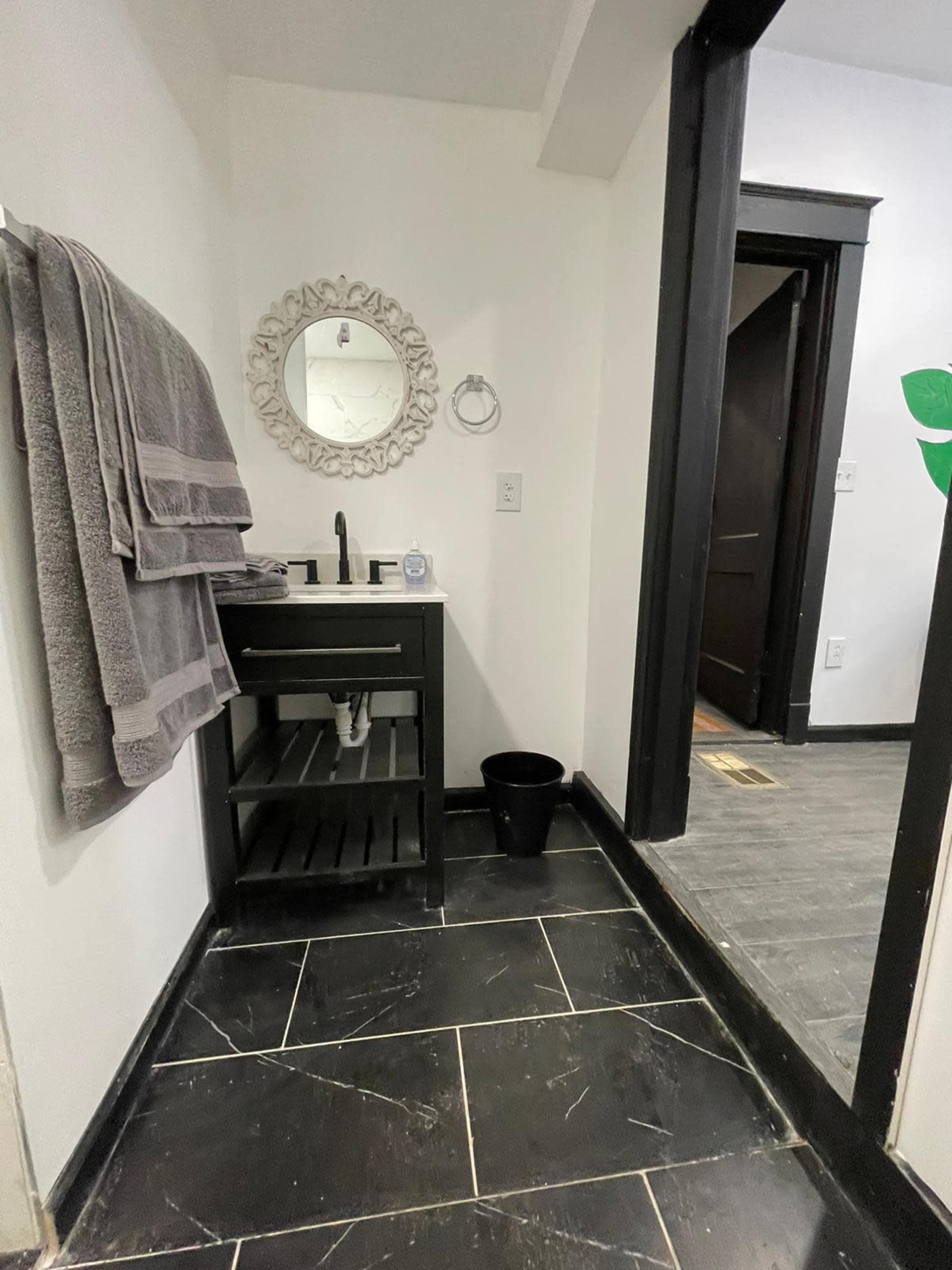 The image shows a bathroom with a dark tiled floor, a black vanity with a mirror above it, and a doorway leading to another room.