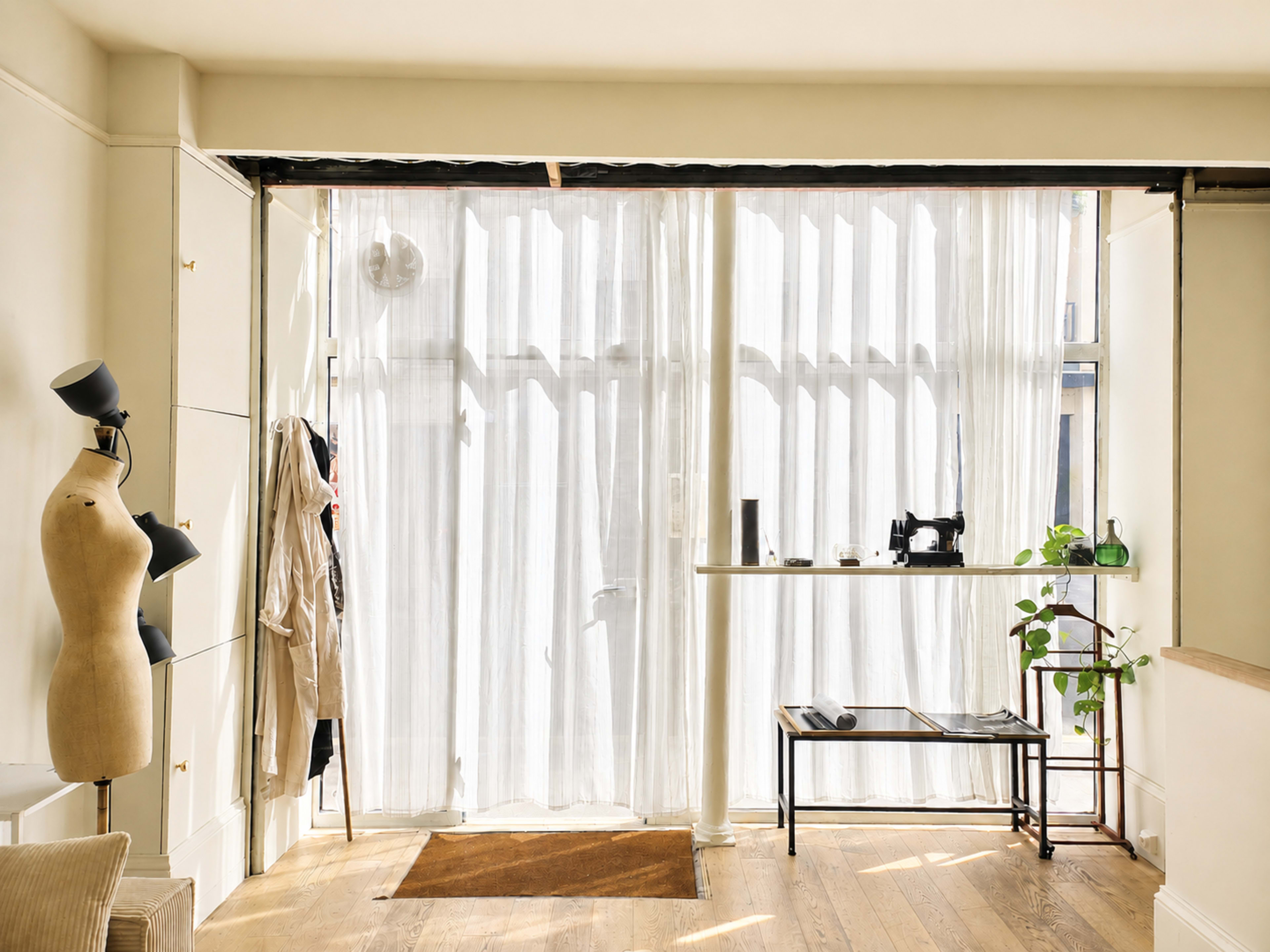 The image shows a well-lit interior space featuring sheer curtains over a window, a mannequin, a small table with a sewing machine, and a potted plant.