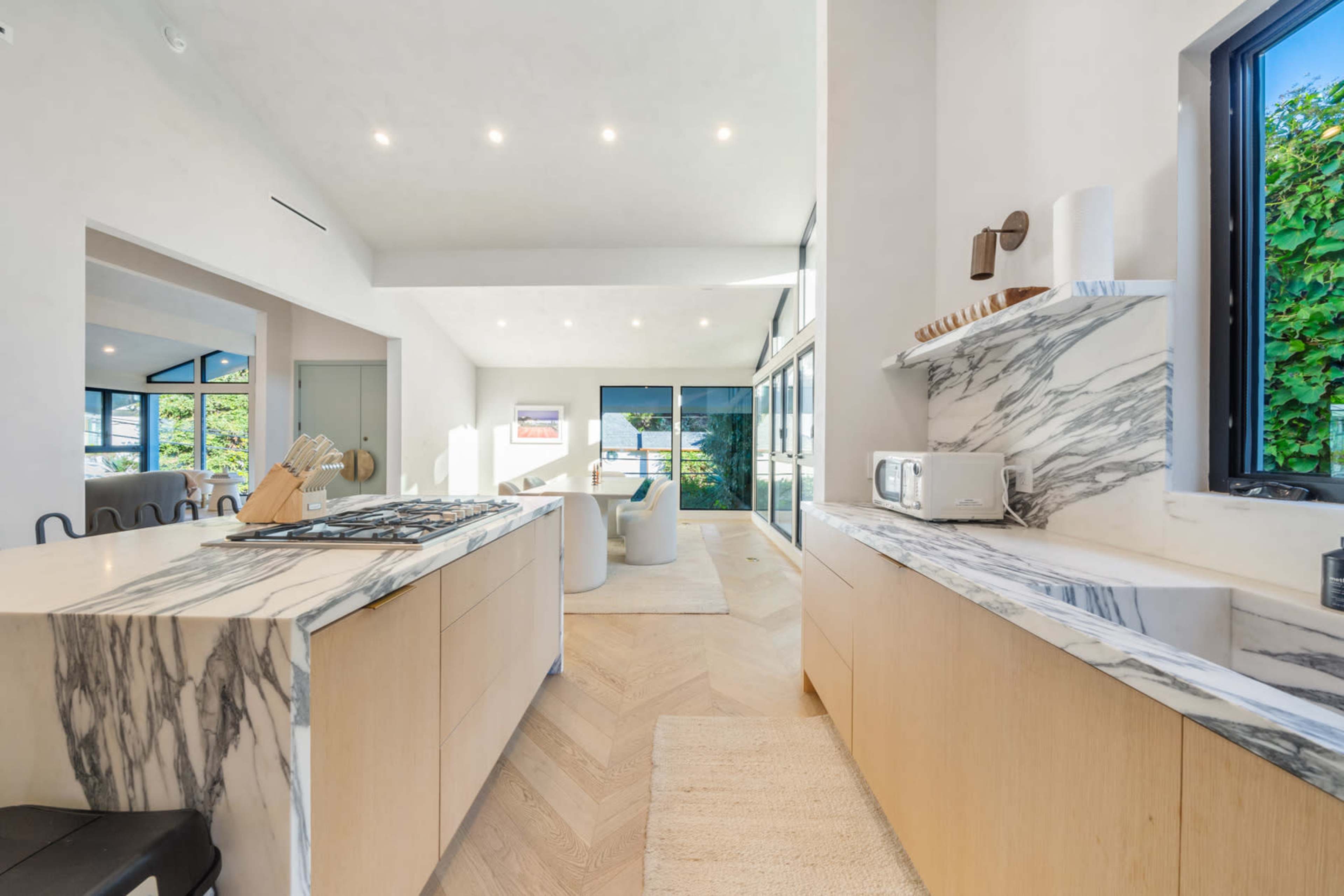The image shows a modern kitchen and living area with marble countertops, light wood cabinetry, and large windows allowing natural light to enter the space.