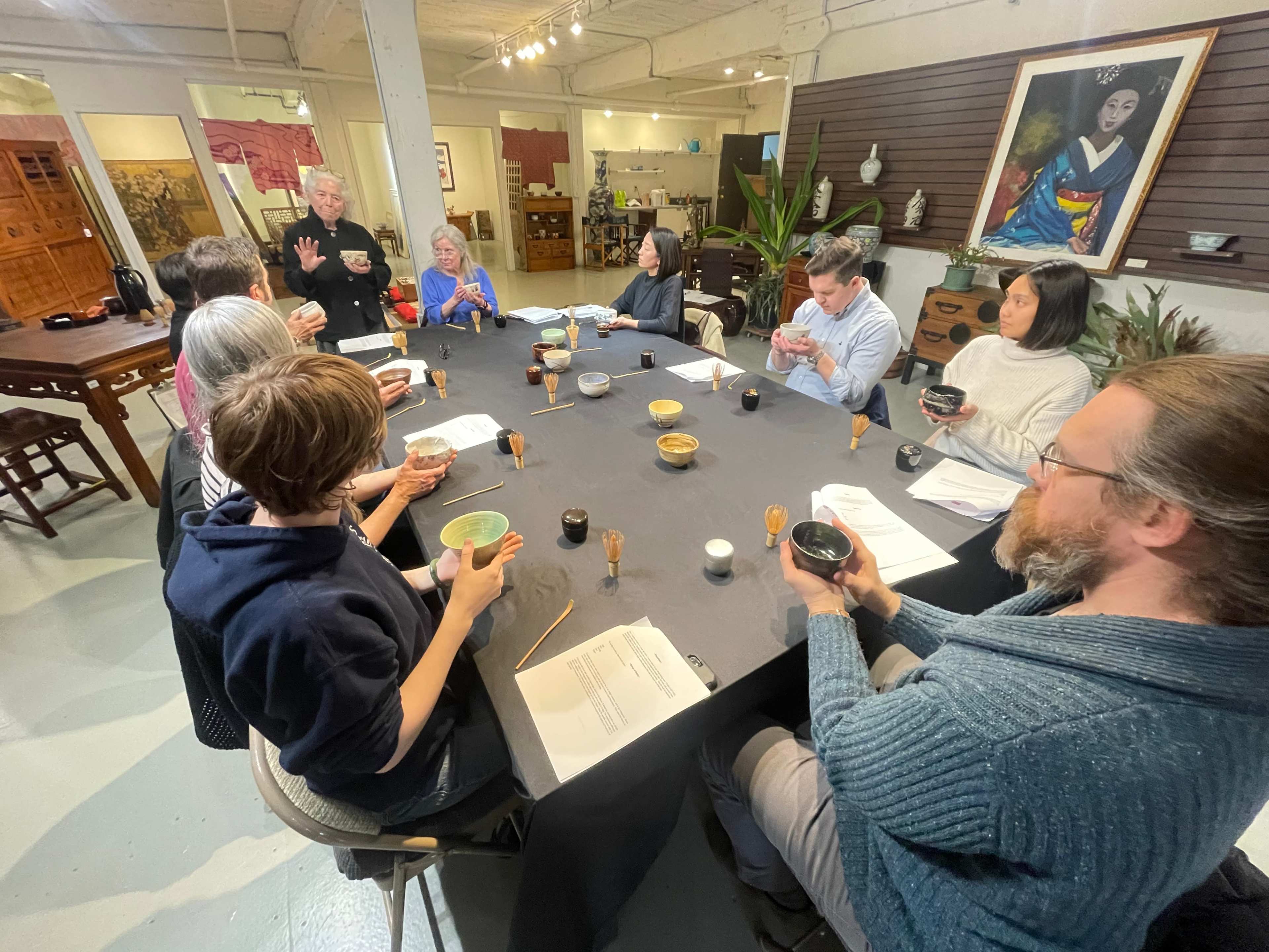 A group of people sits around a large table in a workshop, holding bowls and attentively listening to a speaker.