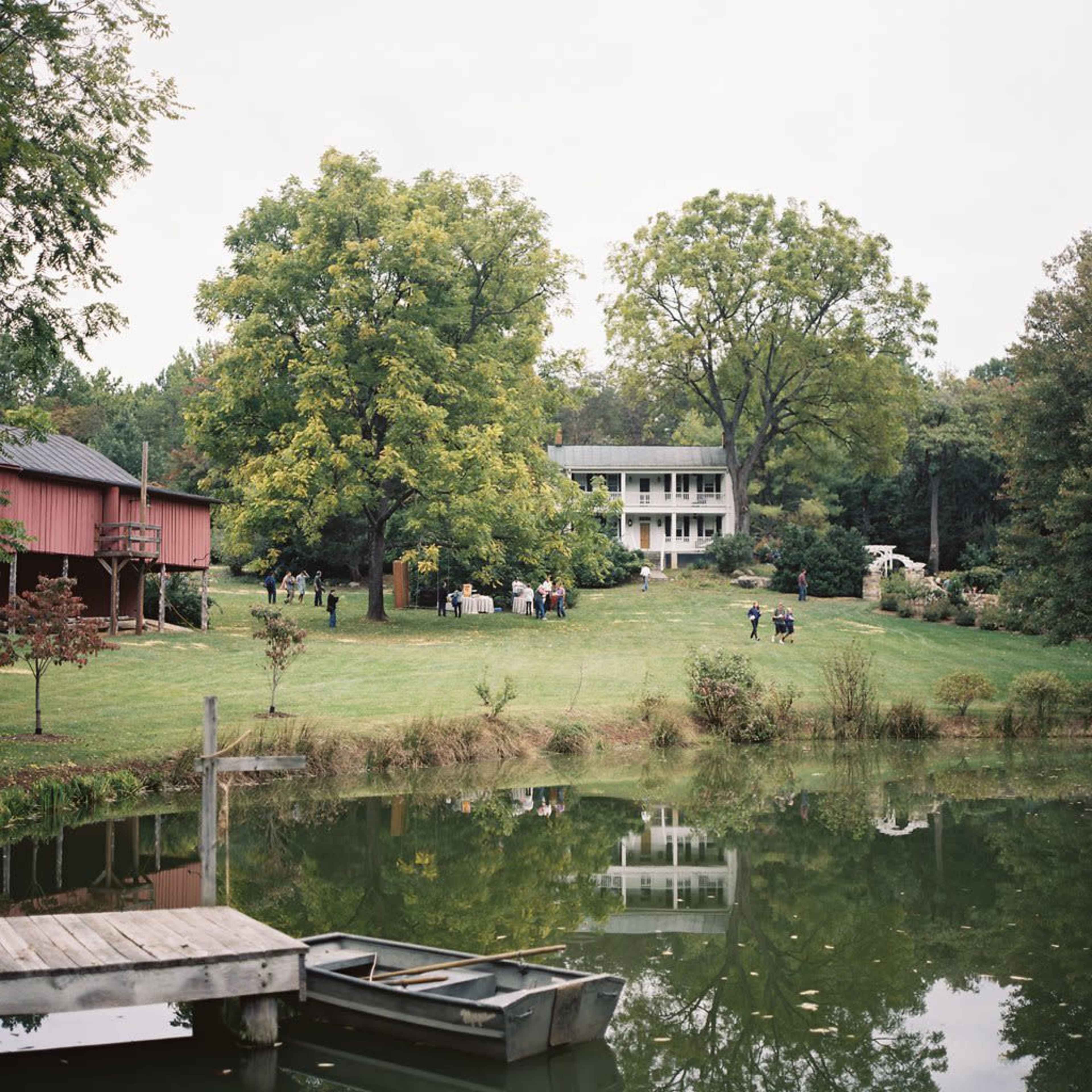 A gathering of people is taking place near a two-story white house by a pond, surrounded by trees and a red barn in the background.