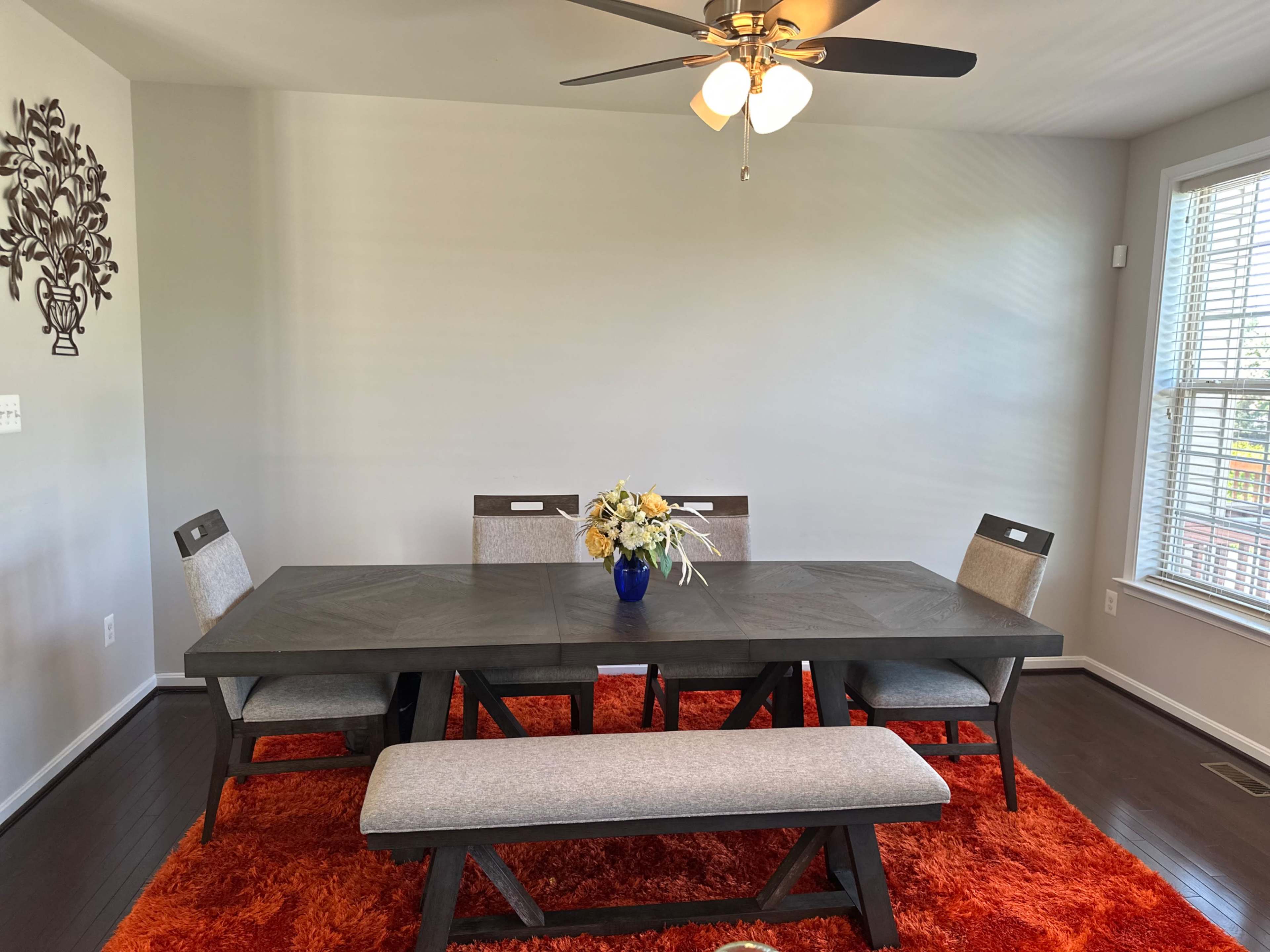 A dining room features a large table surrounded by chairs and a bench, with a vase of flowers in the center and a vibrant orange rug underneath.