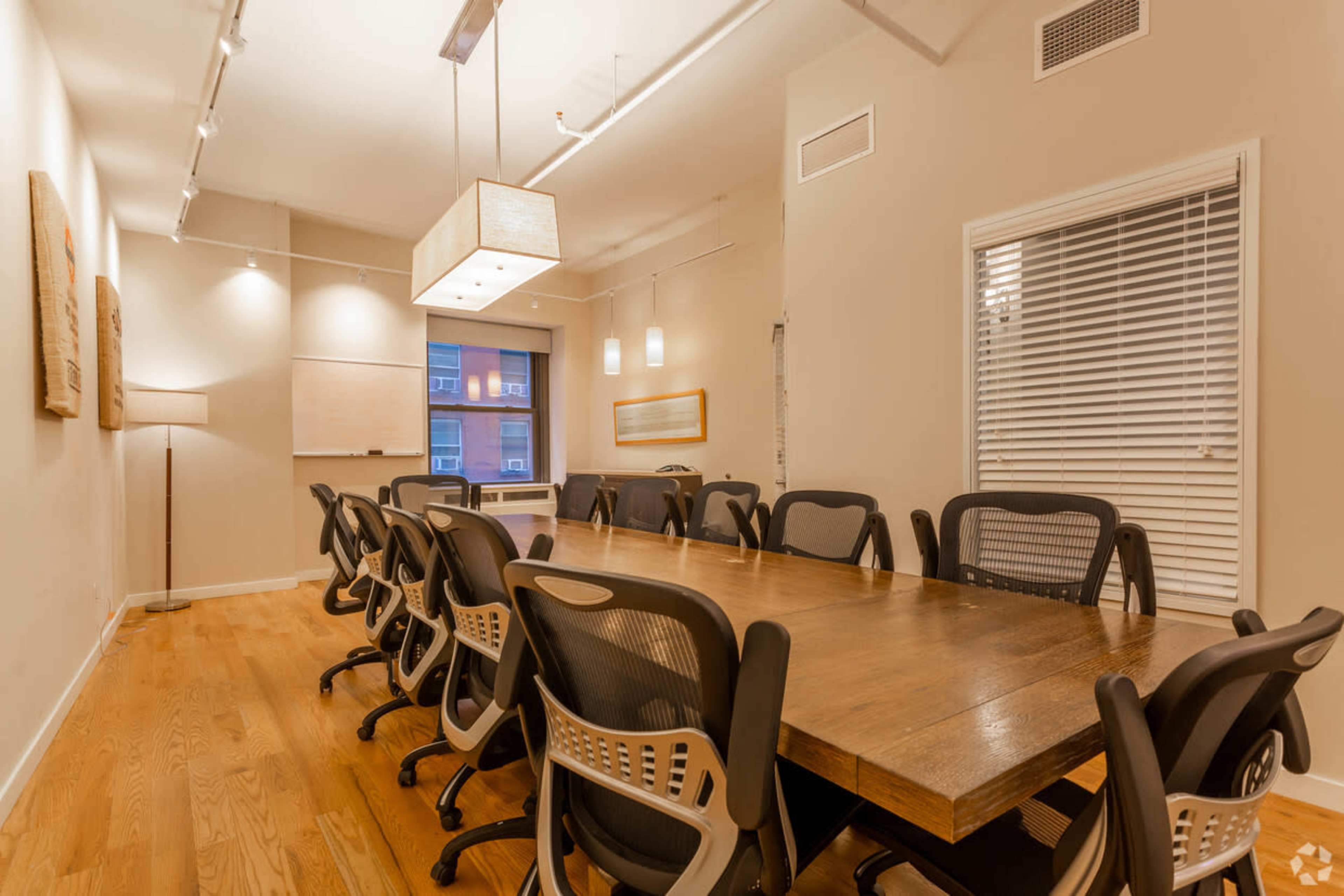 A modern conference room featuring a large wooden table surrounded by several black office chairs, with light fixtures overhead and a window in the background.
