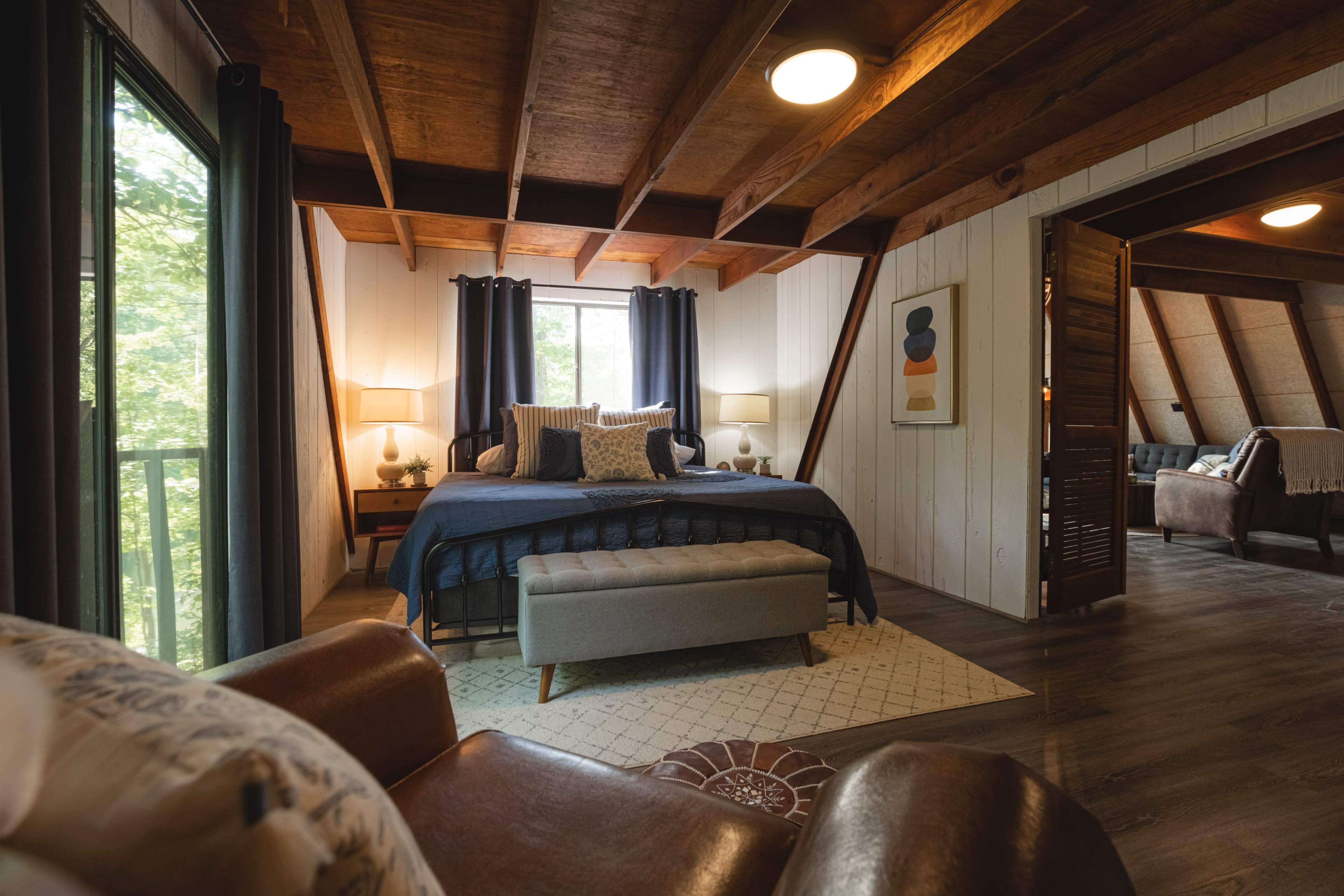 The image shows a cozy attic bedroom with a large bed, a bench at the foot of the bed, and windows allowing natural light to enter, framed by dark curtains.
