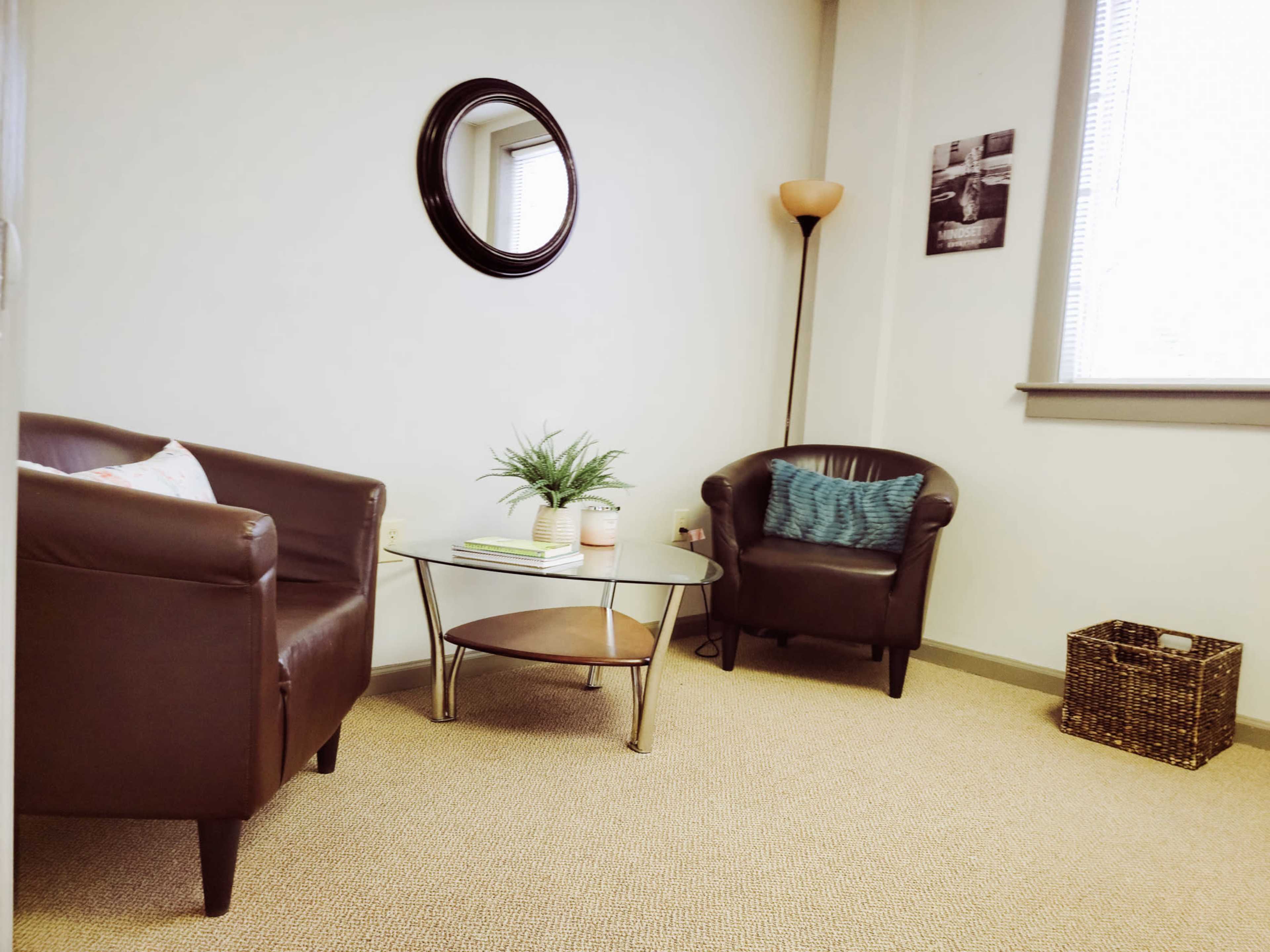 A small office space features two leather armchairs, a round glass coffee table, a potted plant, and a floor lamp, with a mirror and a wall photo in the background.
