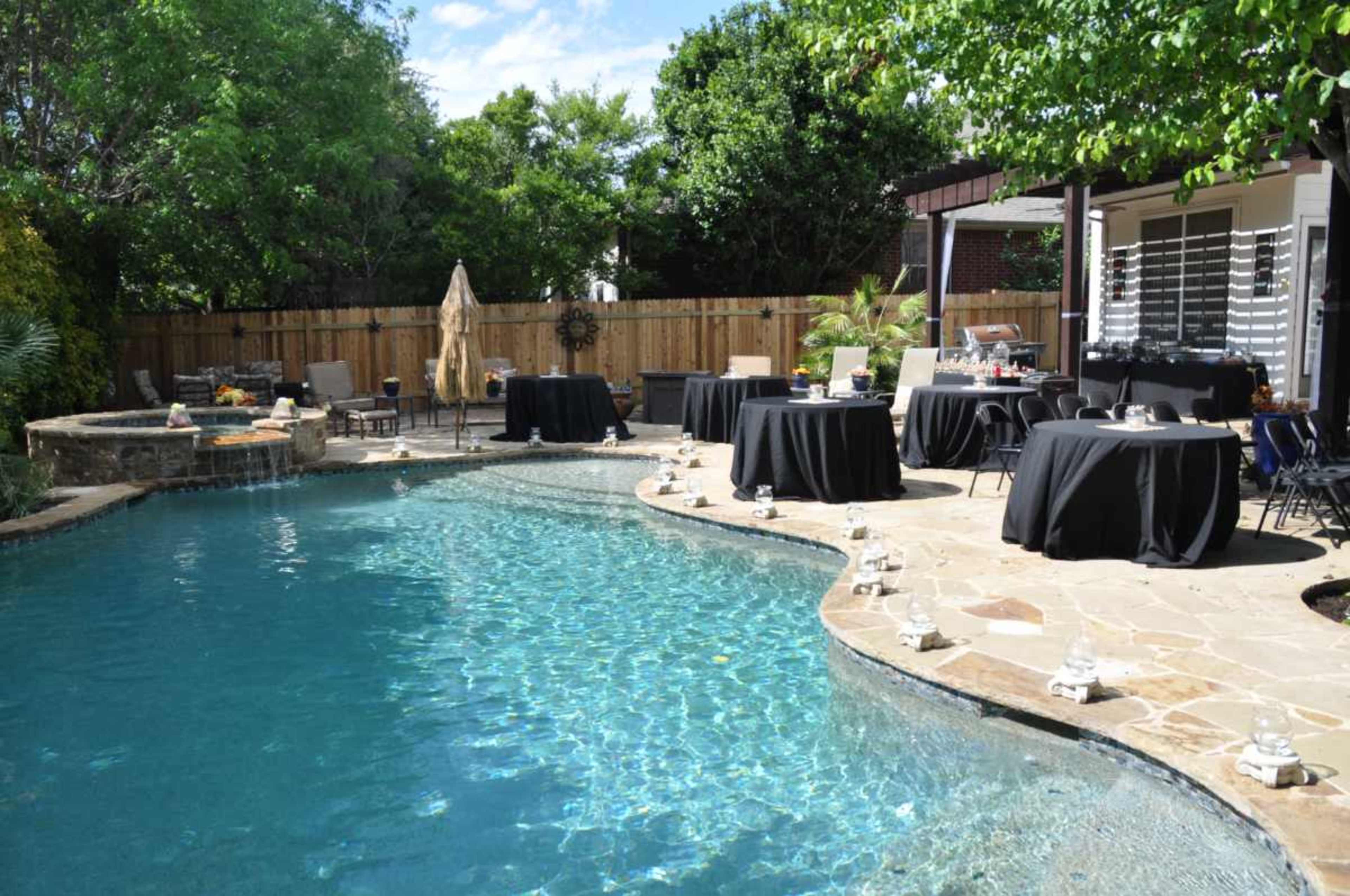 A backyard pool area features a curved pool with stone accents and several tables covered in black cloth arranged around it.