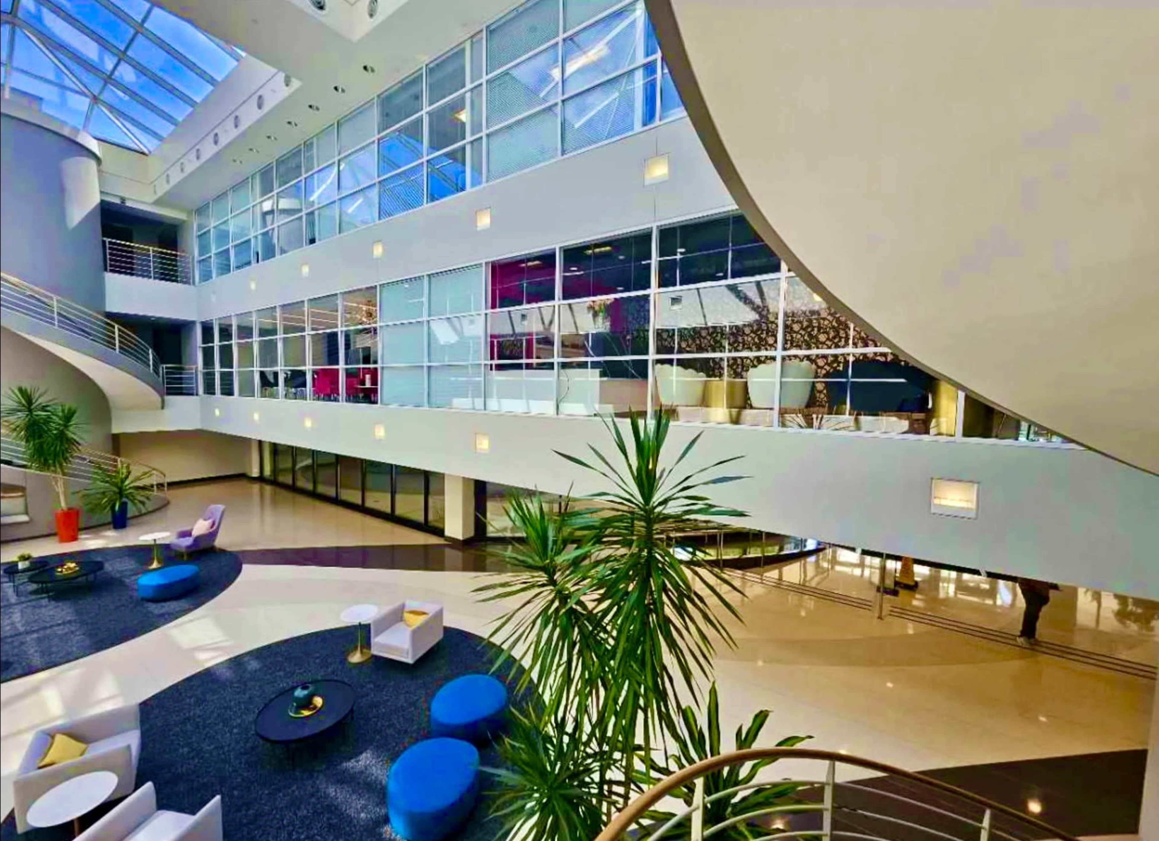 The image shows a modern, spacious lobby with large windows, contemporary furniture, and indoor plants.