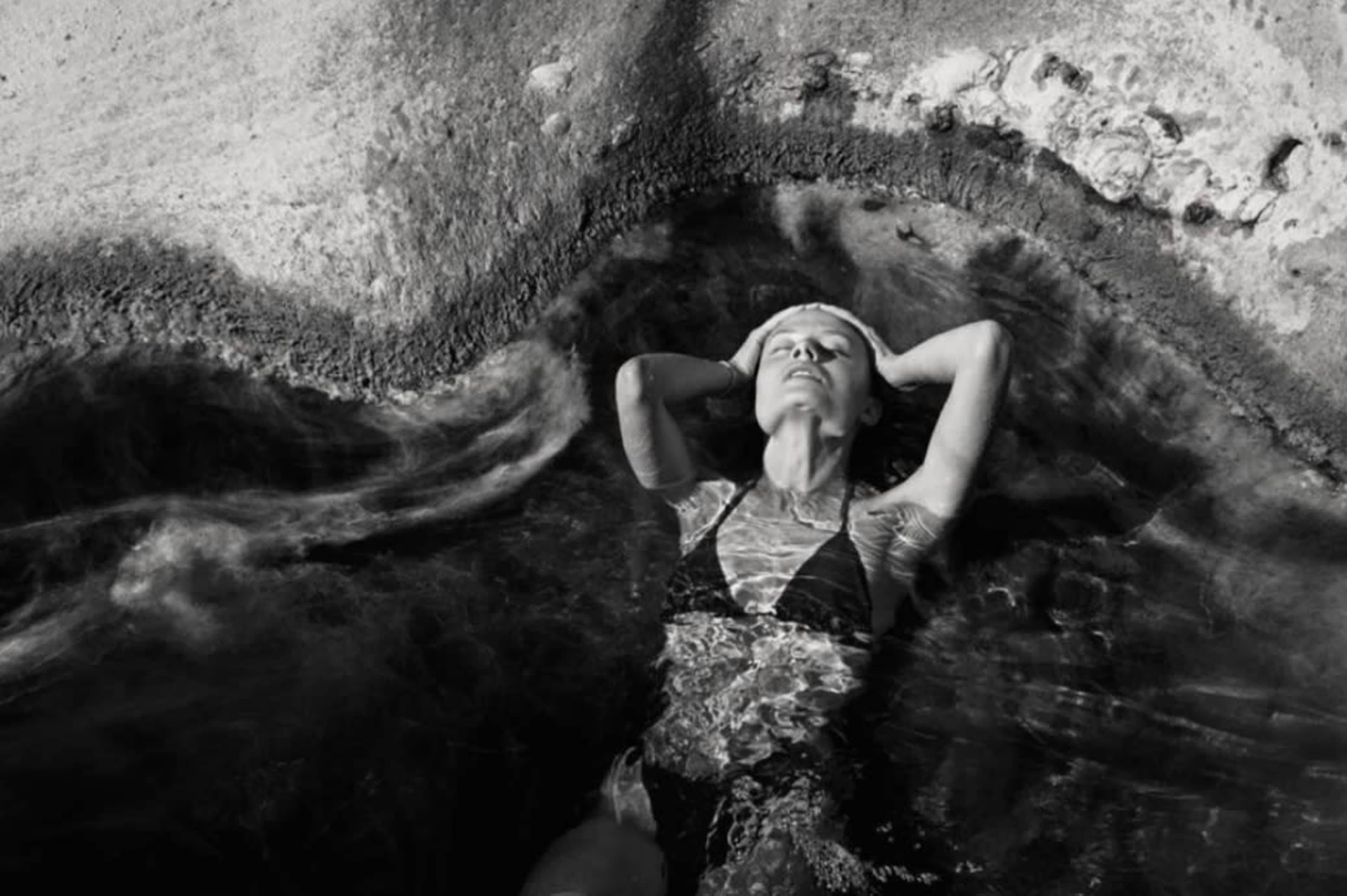 A woman lies on her back in shallow water, surrounded by textured rocky edges.