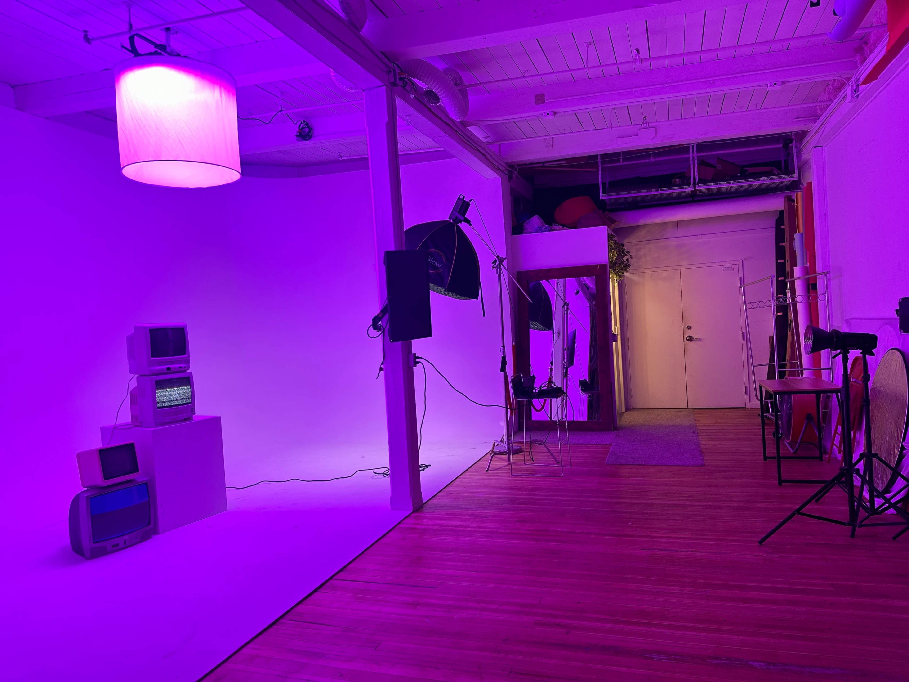 The image shows a photography studio with a purple-lit background, various lighting equipment, an old television set, and a doorway leading to another area.
