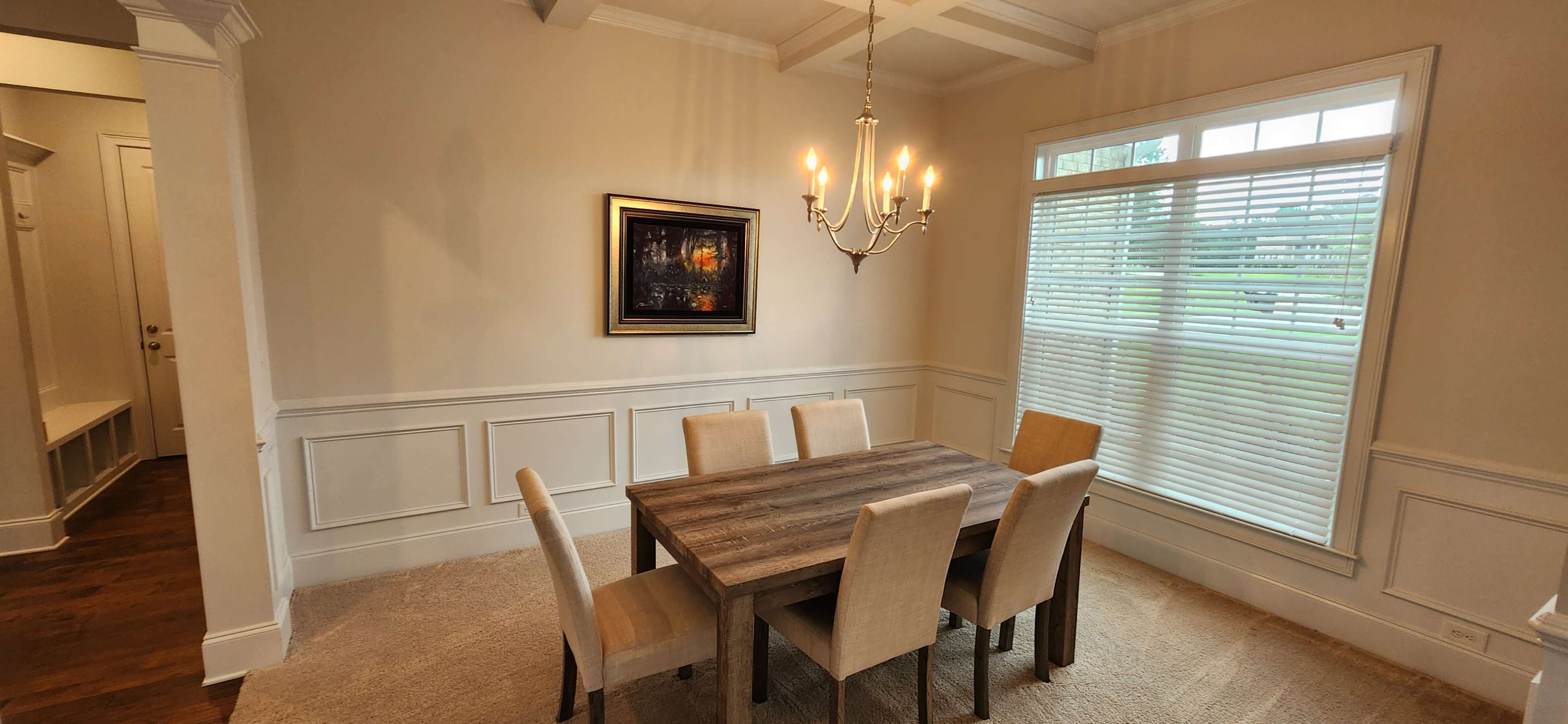 A dining room features a wooden table surrounded by six chairs, with a chandelier overhead and a large window providing natural light.